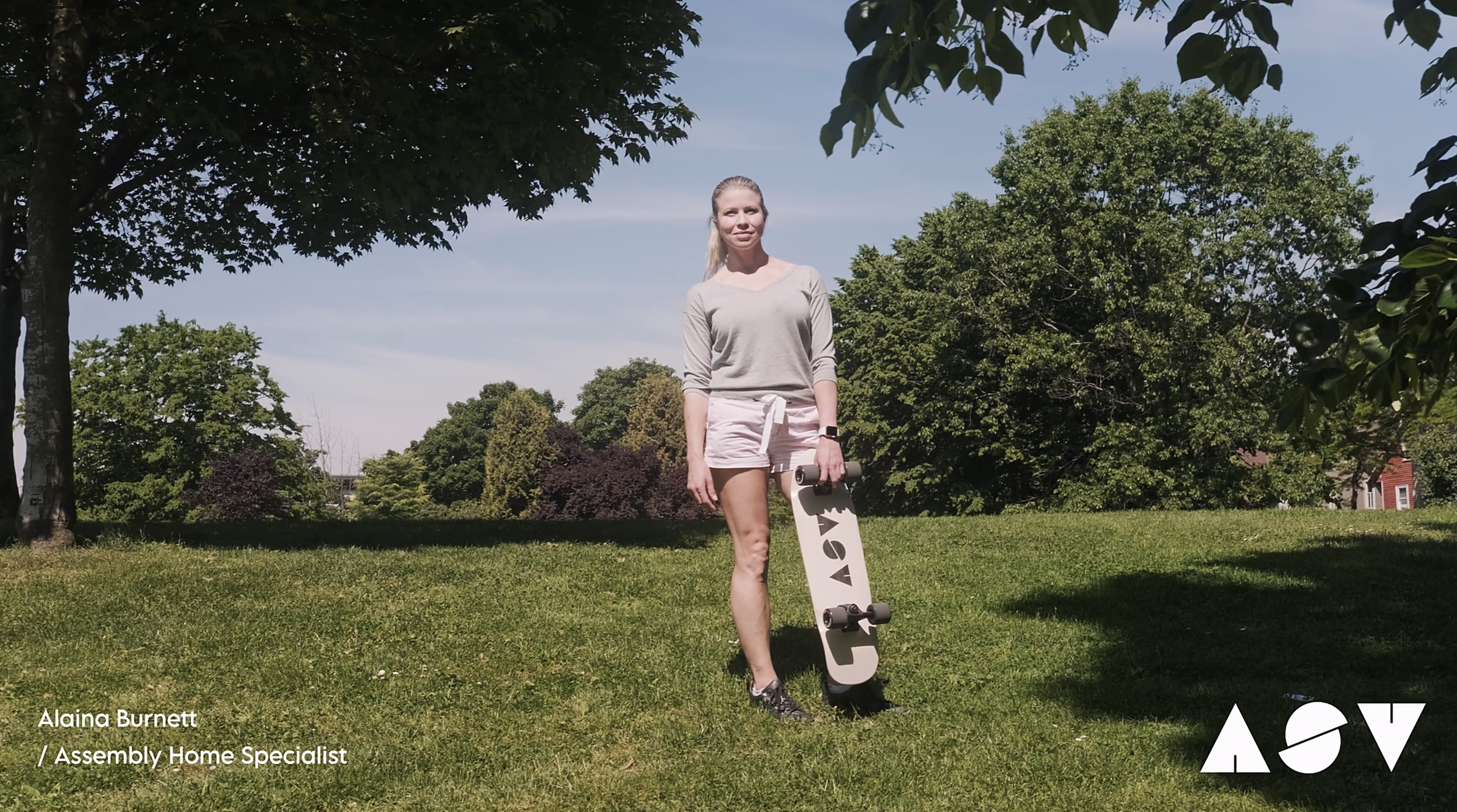 A woman standing on grass holding a skateboard with trees and a blue sky in the background.