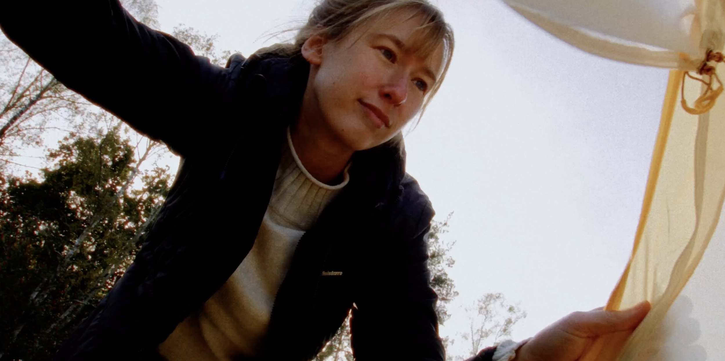 A woman with blonde hair, wearing a cream-colored sweater and black jacket, looks down with a gentle smile while reaching toward the camera. She is outdoors with trees in the background on an overcast day.