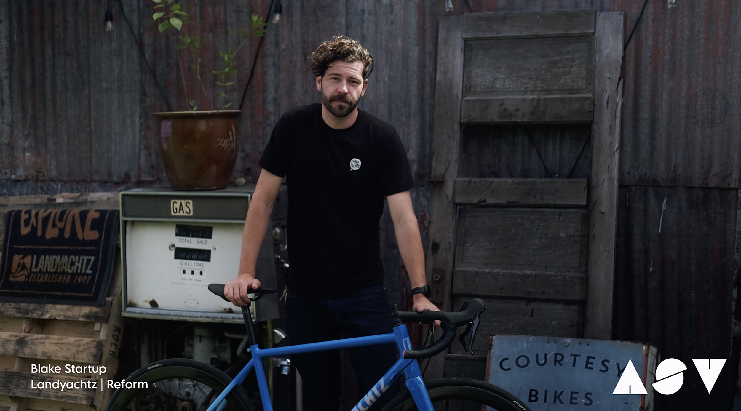 A man with curly hair and a beard, wearing a black T-shirt, standing outdoors with one hand on a blue bicycle handlebar. In the background, there is a wooden fence, a potted plant, a white gas pump, and a sign that says 'Courtes' and 'Bikes'.