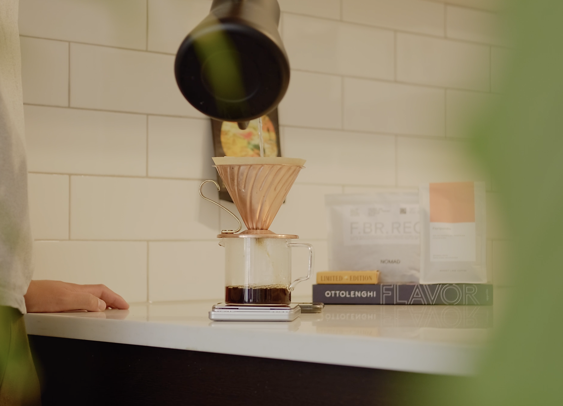 A Chemex coffee maker with a paper filter, placed on a kitchen counter, with a background of books and constrained by a hand resting on the counter.