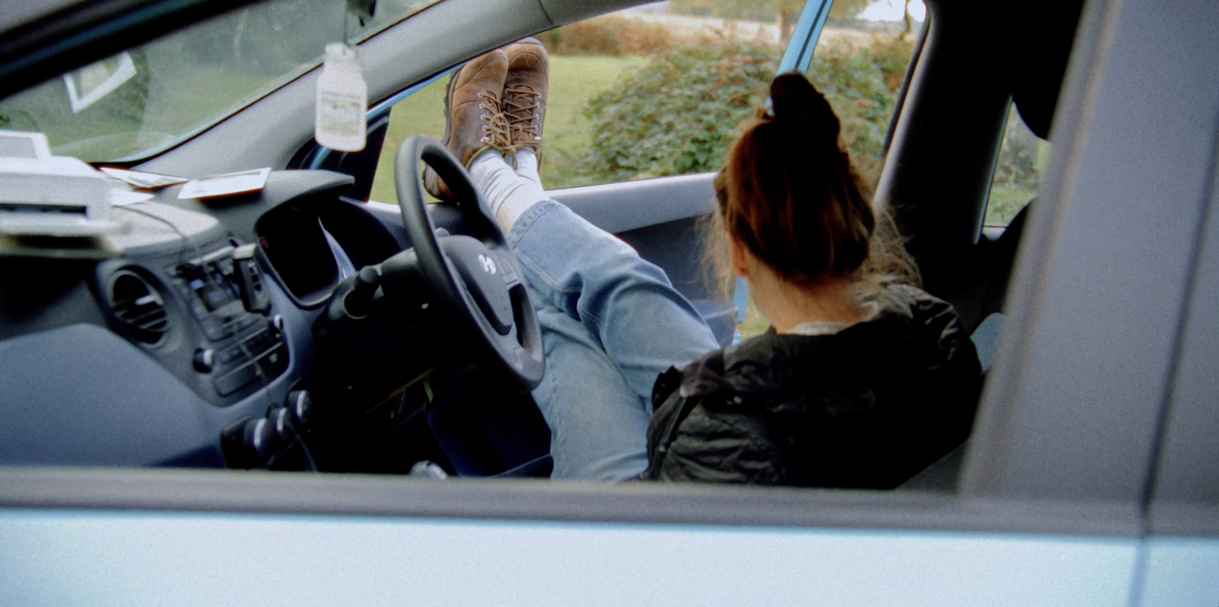 Person with brown hair tied back, sitting in the passenger seat of a vehicle, looking outside through the windshield. The person's shoes are propped up on the dashboard. The vehicle's dashboard with various controls and a small white object hanging from the rearview mirror are visible.