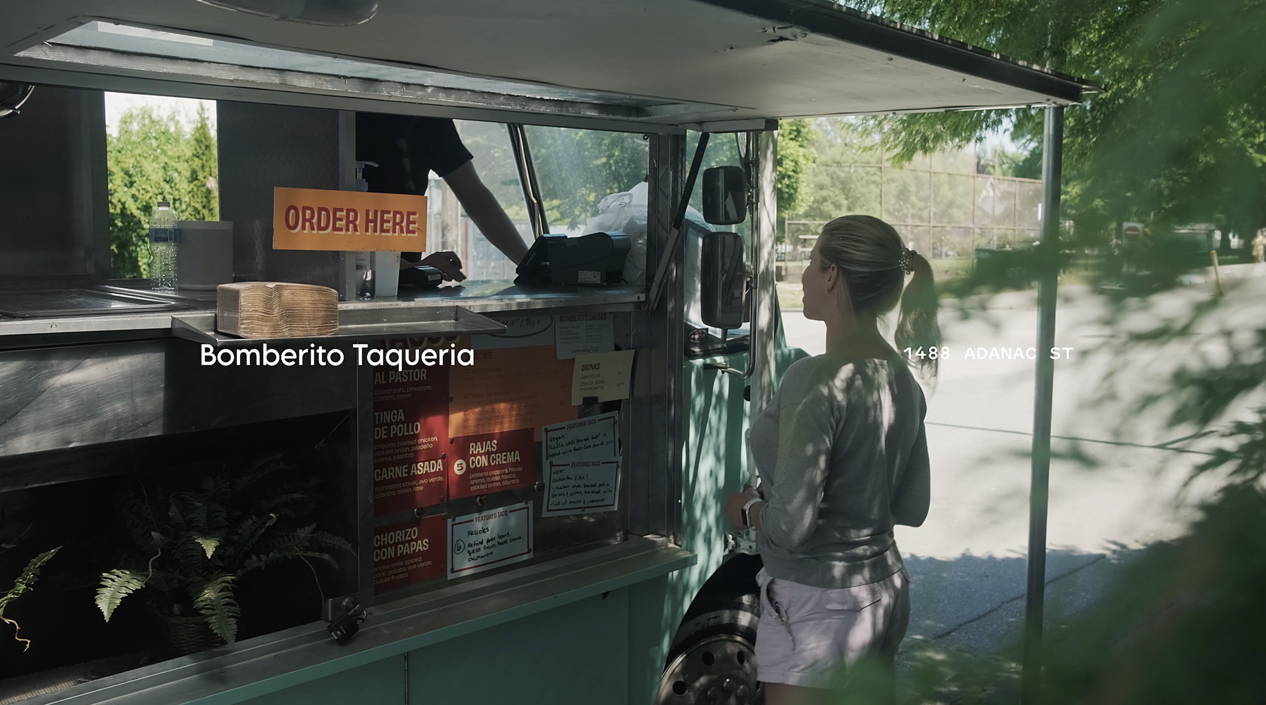 A woman placing an order at a taco food truck named Bomberito Taqueria during daytime. The truck has a sign that says 'ORDER HERE' and a menu on the side. The setting is outdoors with trees and a tennis court in the background.