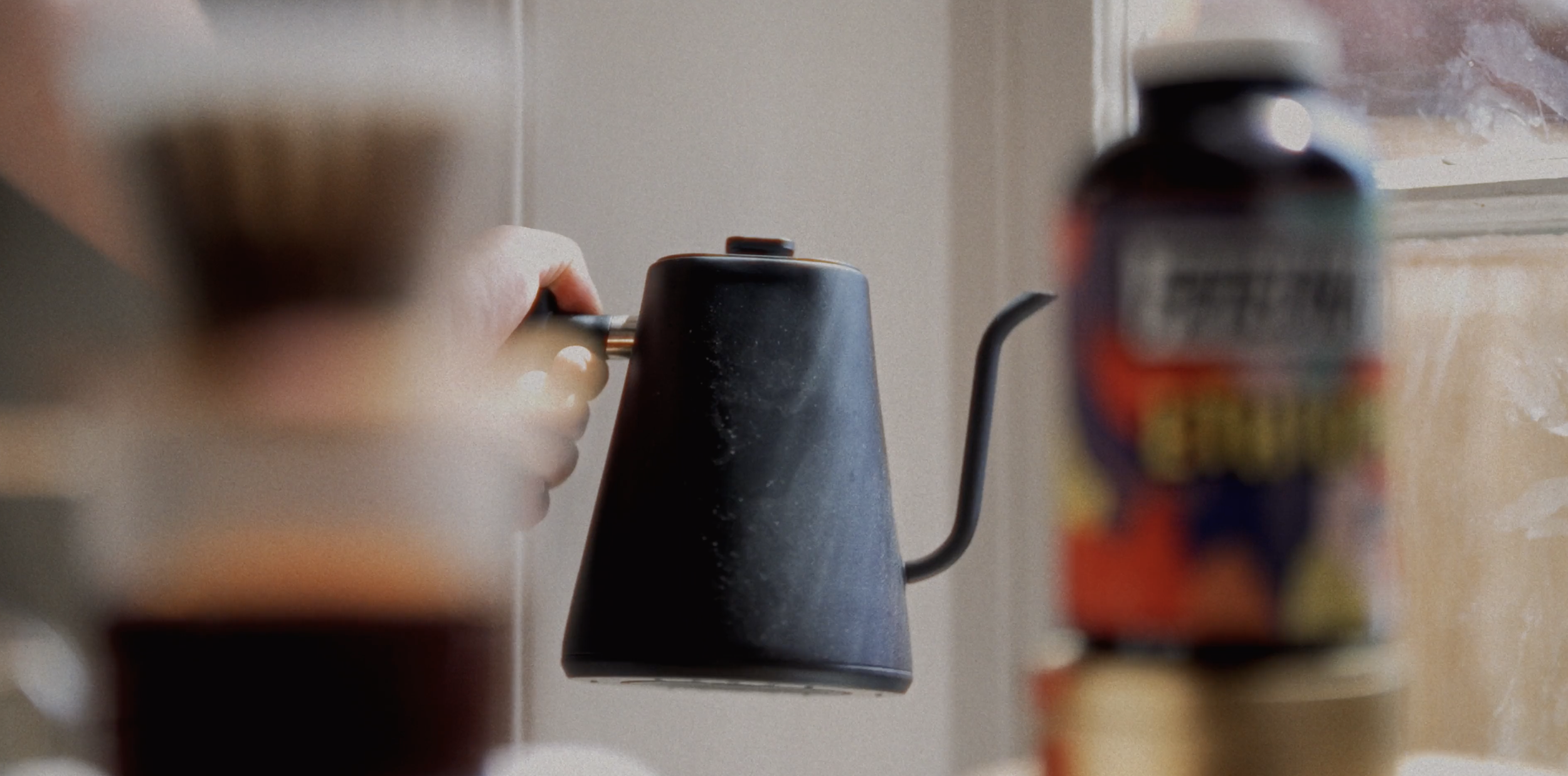 A person holding a black gooseneck kettle in a kitchen with a beige wall, with some blurry objects in the foreground.