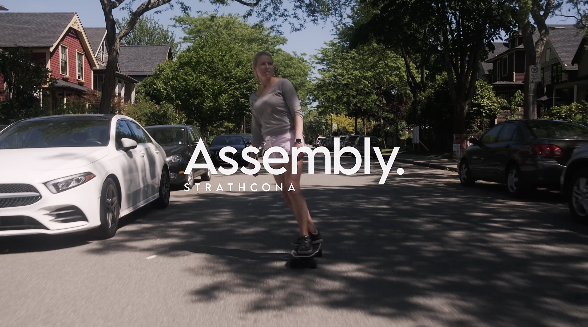 Person skateboarding across a city street with parked cars and houses in the background, with text overlay reading 'Assembly. STRATHCONA'.