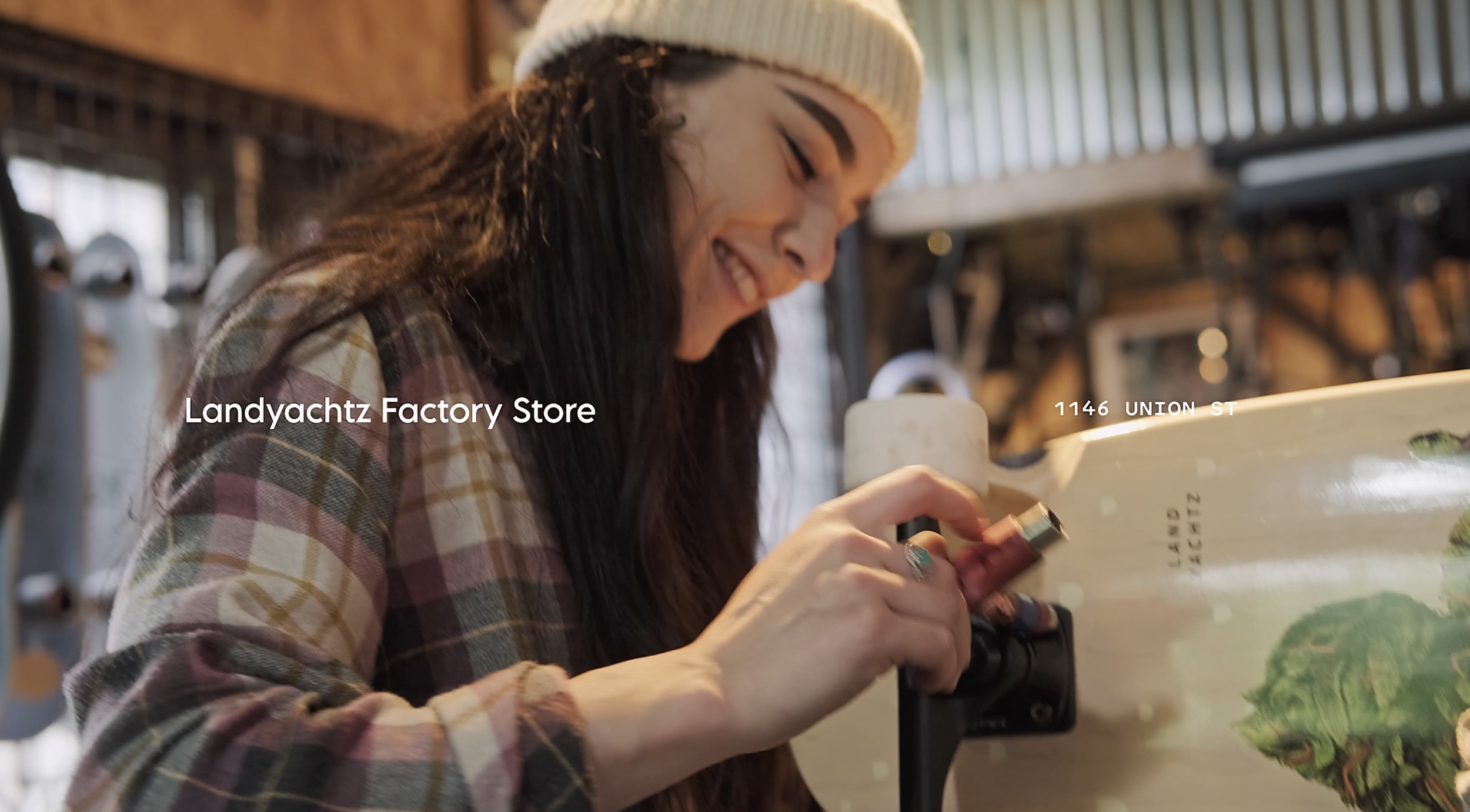 A woman in a beige beanie and plaid shirt smiling and looking at her phone inside a store with wooden walls. The store is called Landyachtz Factory Store and is located at 1146 Union Street.