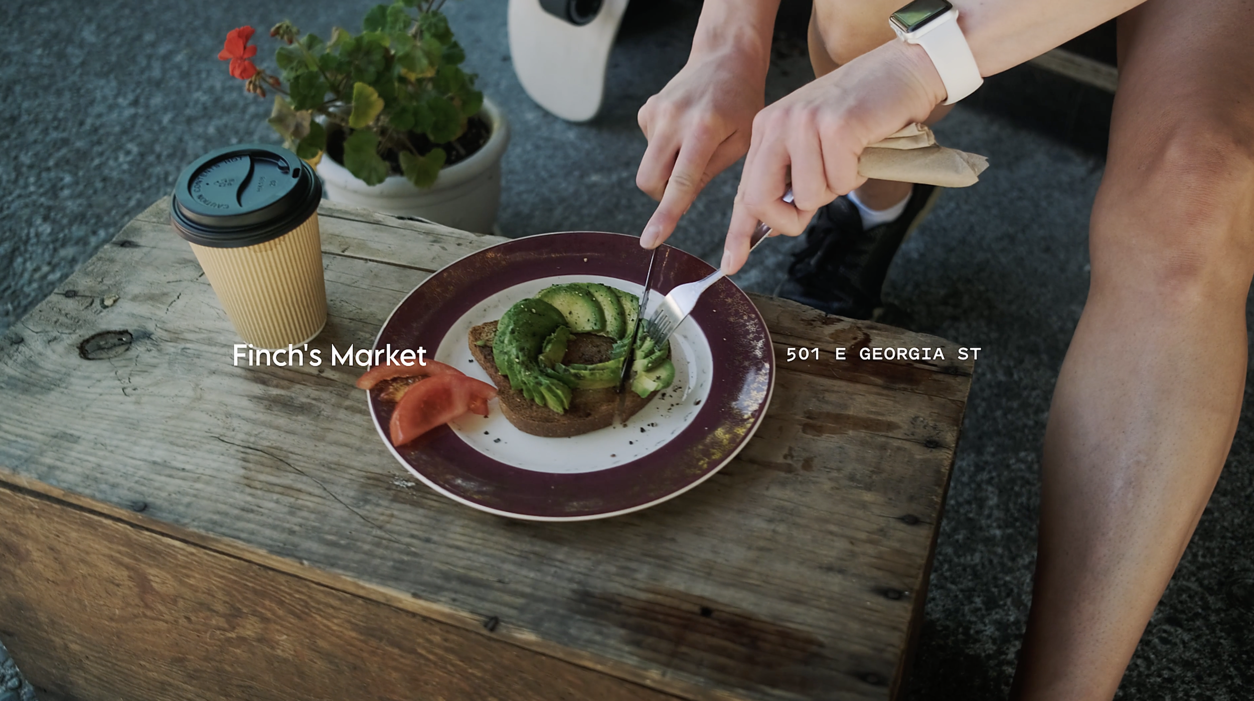 A top view of a person slicing avocado on a plate with toast, tomato, and other toppings, next to a coffee cup and a small potted plant on a rustic wooden table.