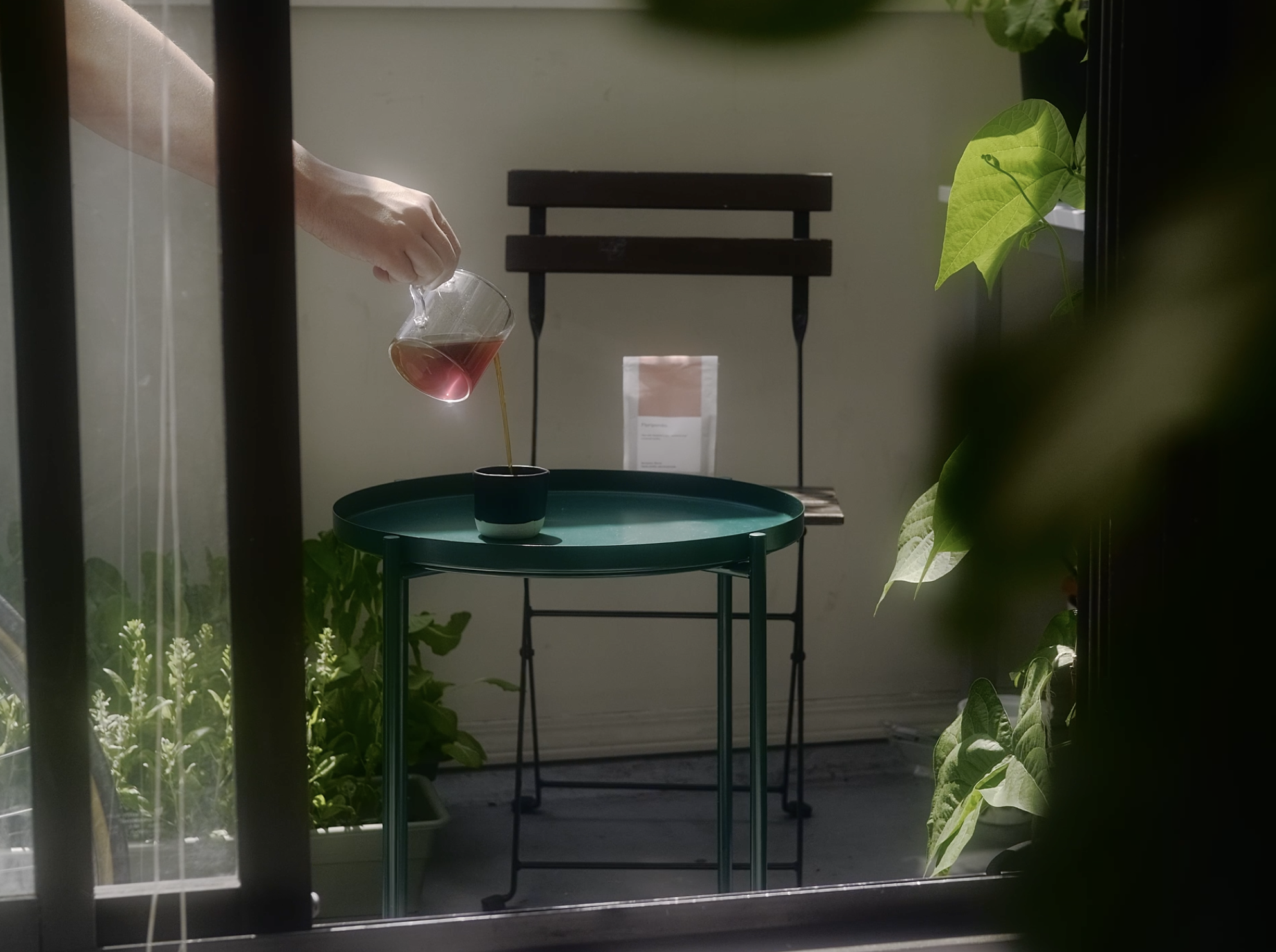 A person's hand pouring a pink liquid into a black cup on a green table, seen through a window with greenery on the sides.