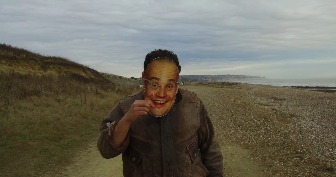 A man walks along a coastal path wearing an Al Murray mask. A still from short film, The Mask.