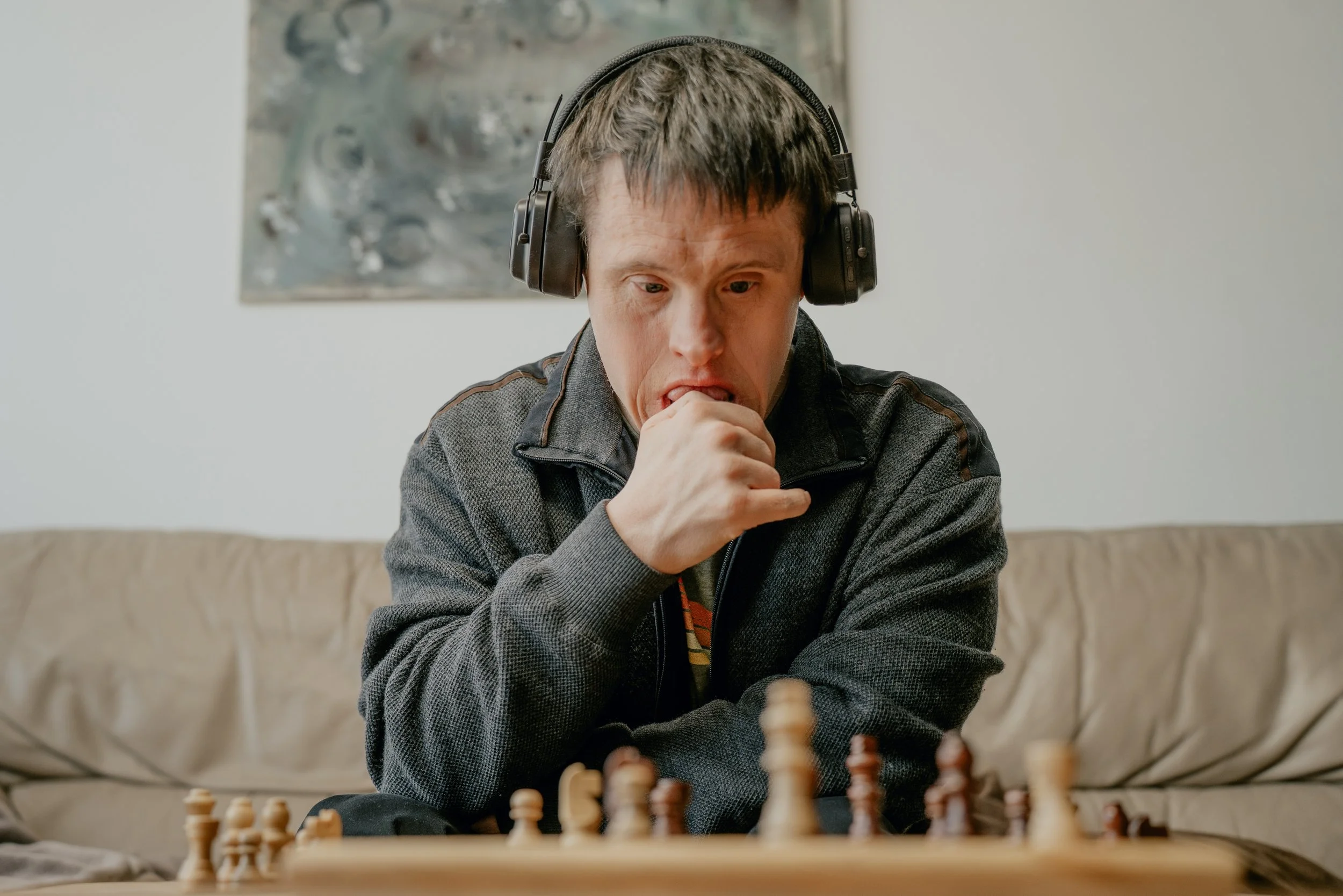 A Downs Syndrome man wearing headphones sitting down looking at a chess board with one hand near his mouth. He looks like he is concentrating.