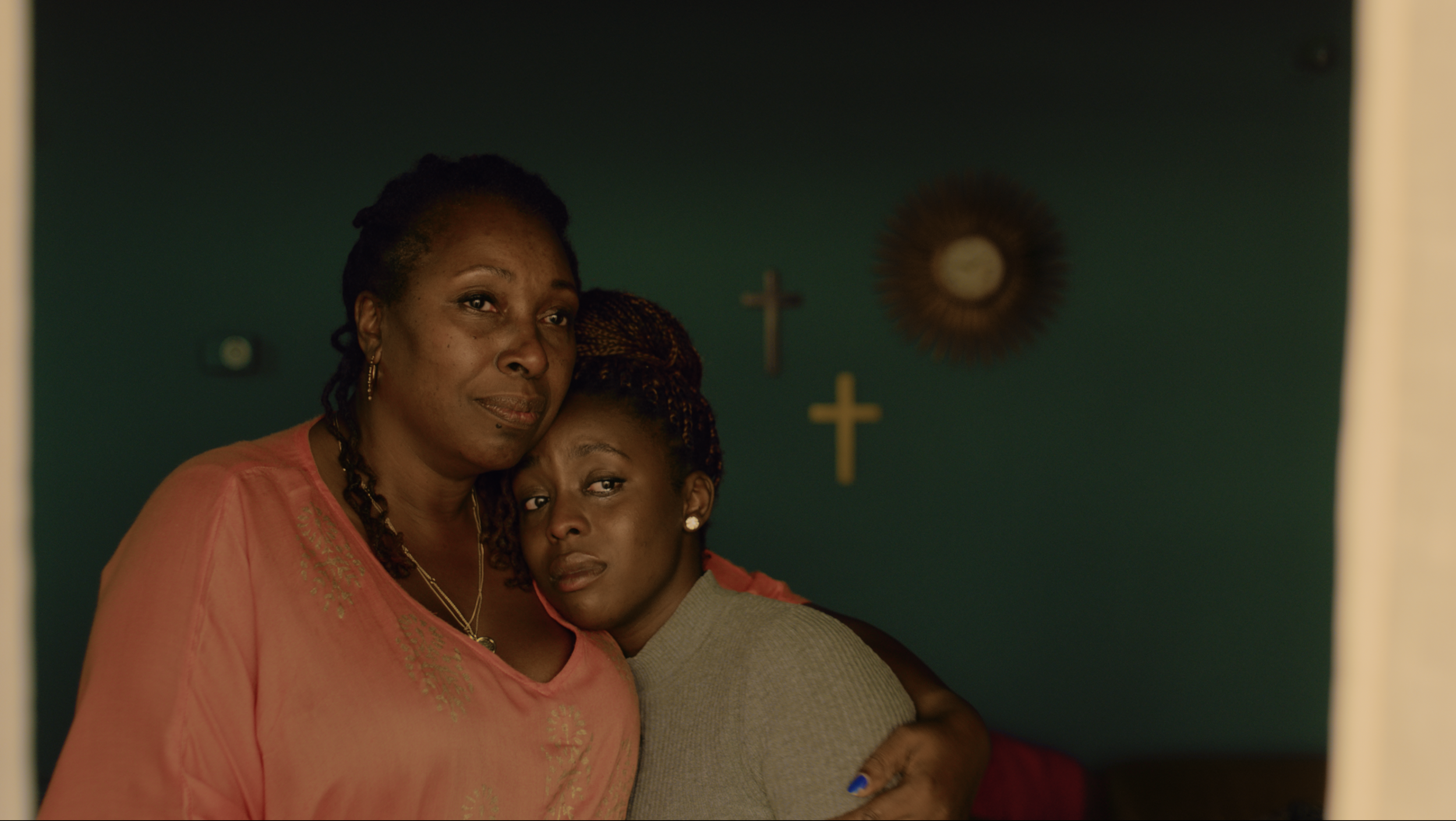 A film still from ‘The Call’ showing the two main characters, a mother and daughter in an embrace. The daughter is tearful and the mother is gazing ahead as if looking out of the window, with a gentle smile on her face.