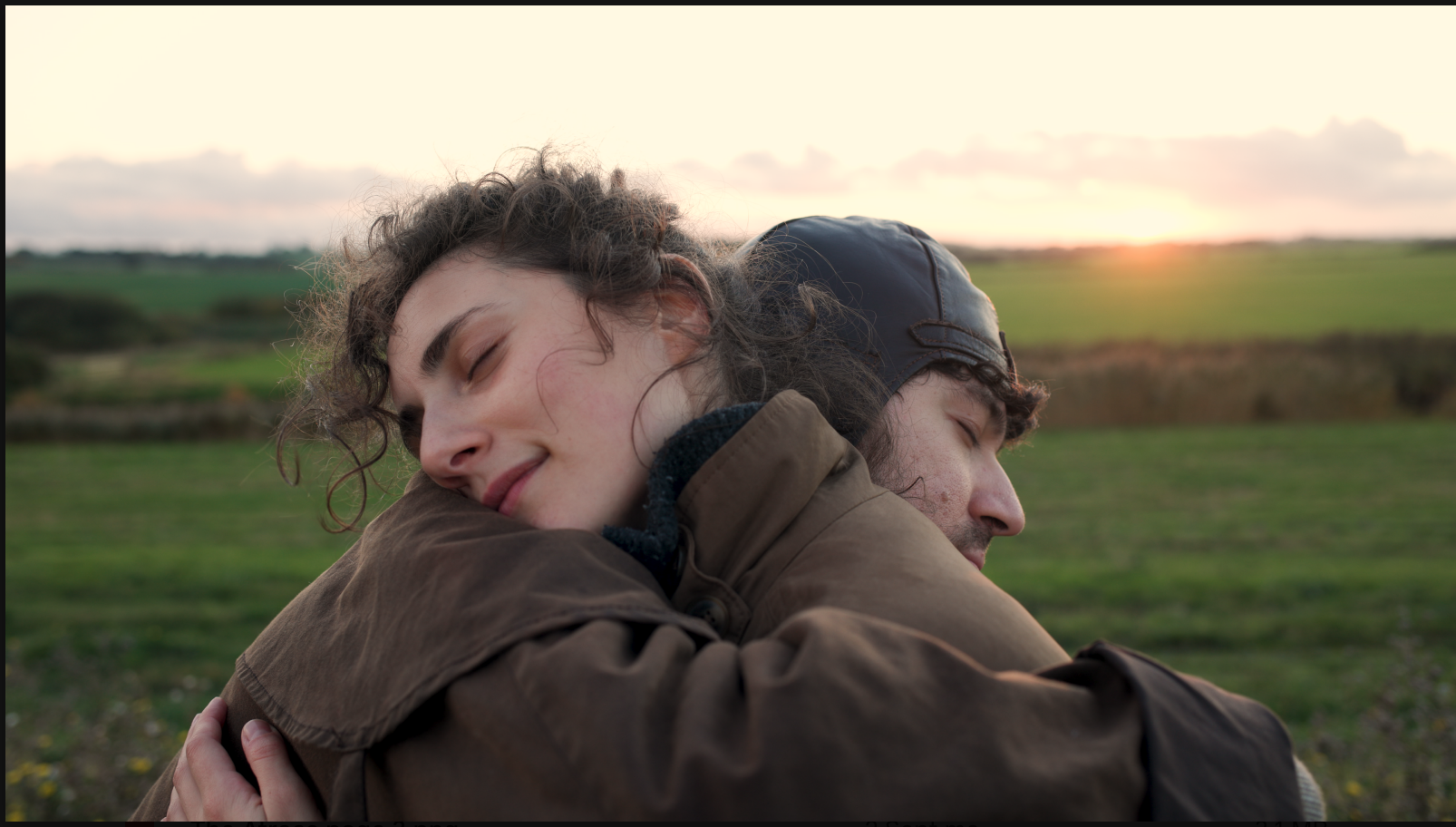 A man and a woman hug shoulder to shoulder. They are in a field and sunset is just about to happen.He is wearing an old fashioned leather motorcycle hat.