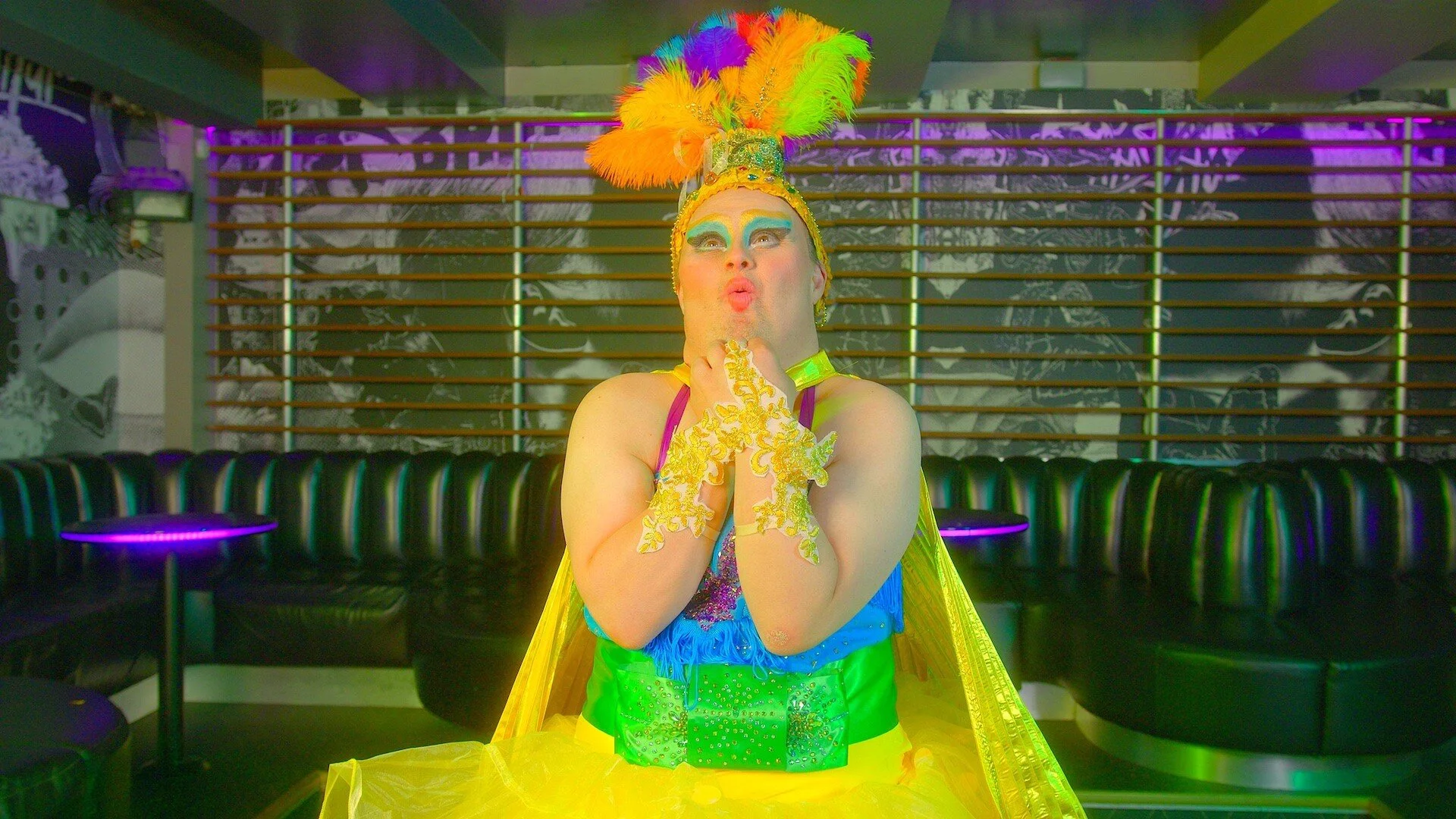 A man dressed in a colourful dress, bold bright makeup and a feathered head dress. His hands are clasped underneath his face.