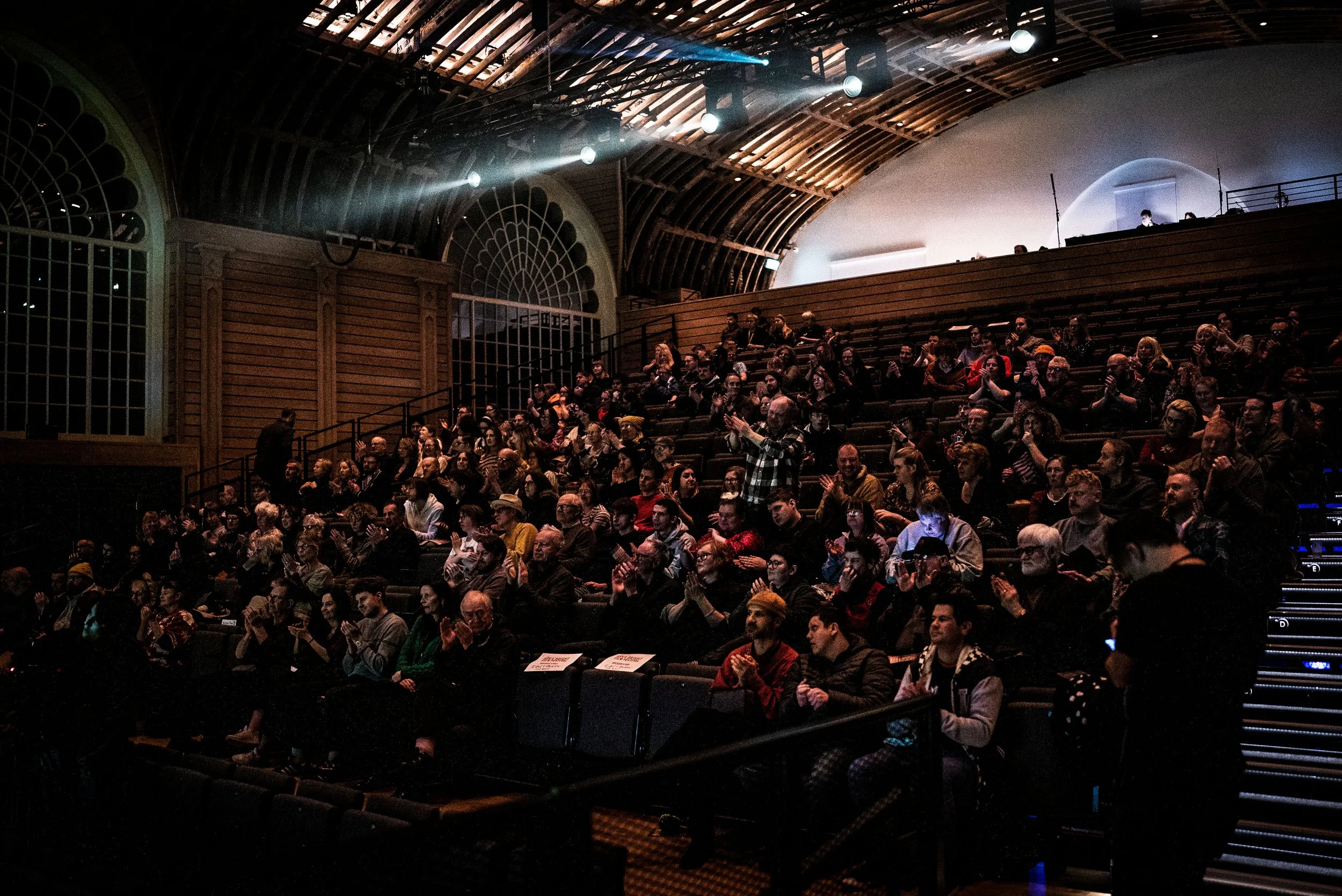 Oska Bright audience members are sat on the bleachers seats in the Corn Exchange at Brighton Dome. The audience members are clapping.