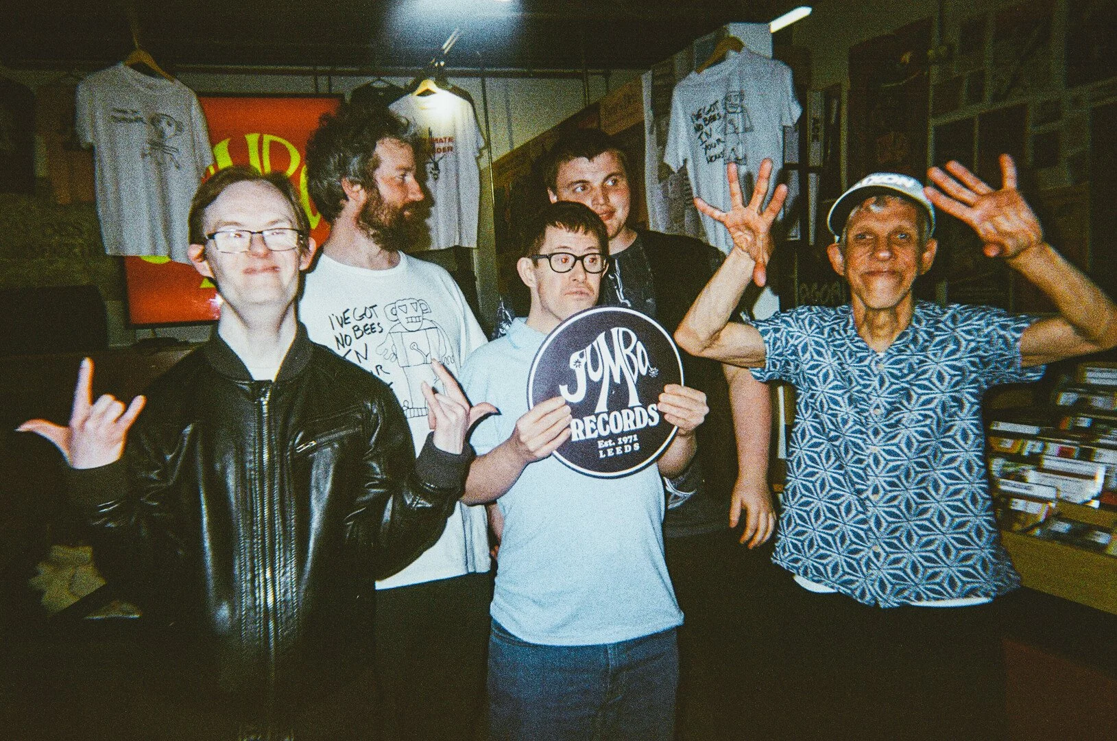 five men stood in a group, three in front 2 behind. the central person is holding a circular sign saying 'jumbo records'.