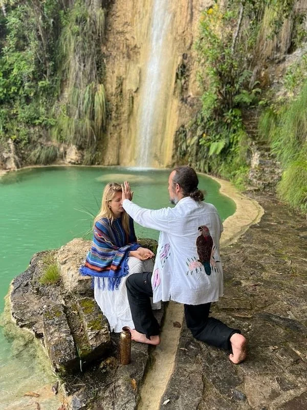 Shamanic teacher Alku, standing in front of a participant during a Munay-Ki rite of passage in nature, epic waterfall setting with turquoise water, Aynua Amazonas, Peru