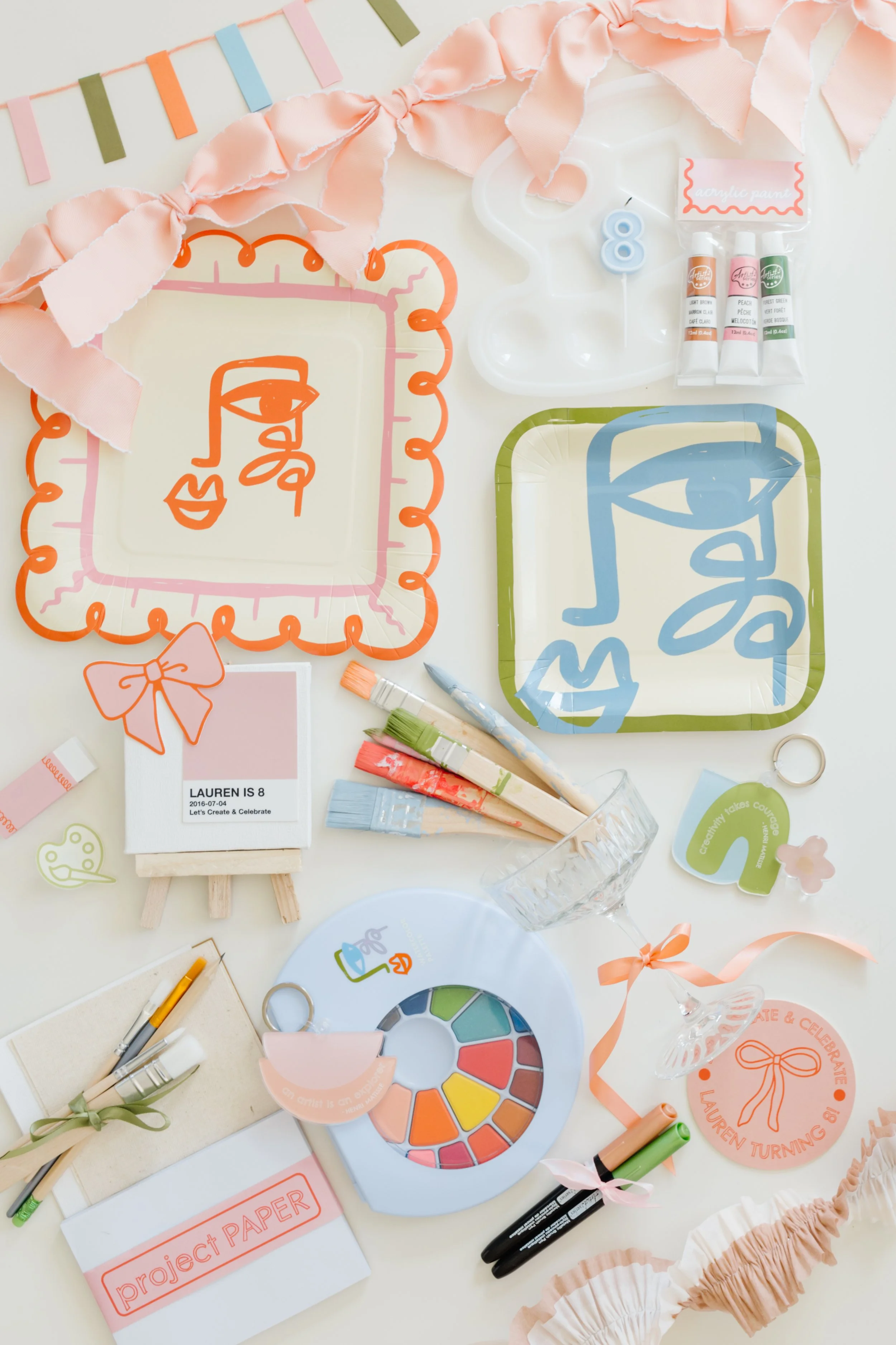 Decorative table setup for a birthday party with pastel-colored plates, banners, and art supplies including paint, brushes, and markers. There are birthday-themed signs, a pink frilled table runner, and a name card indicating it is for an eight-year-old girl named Lauren.
