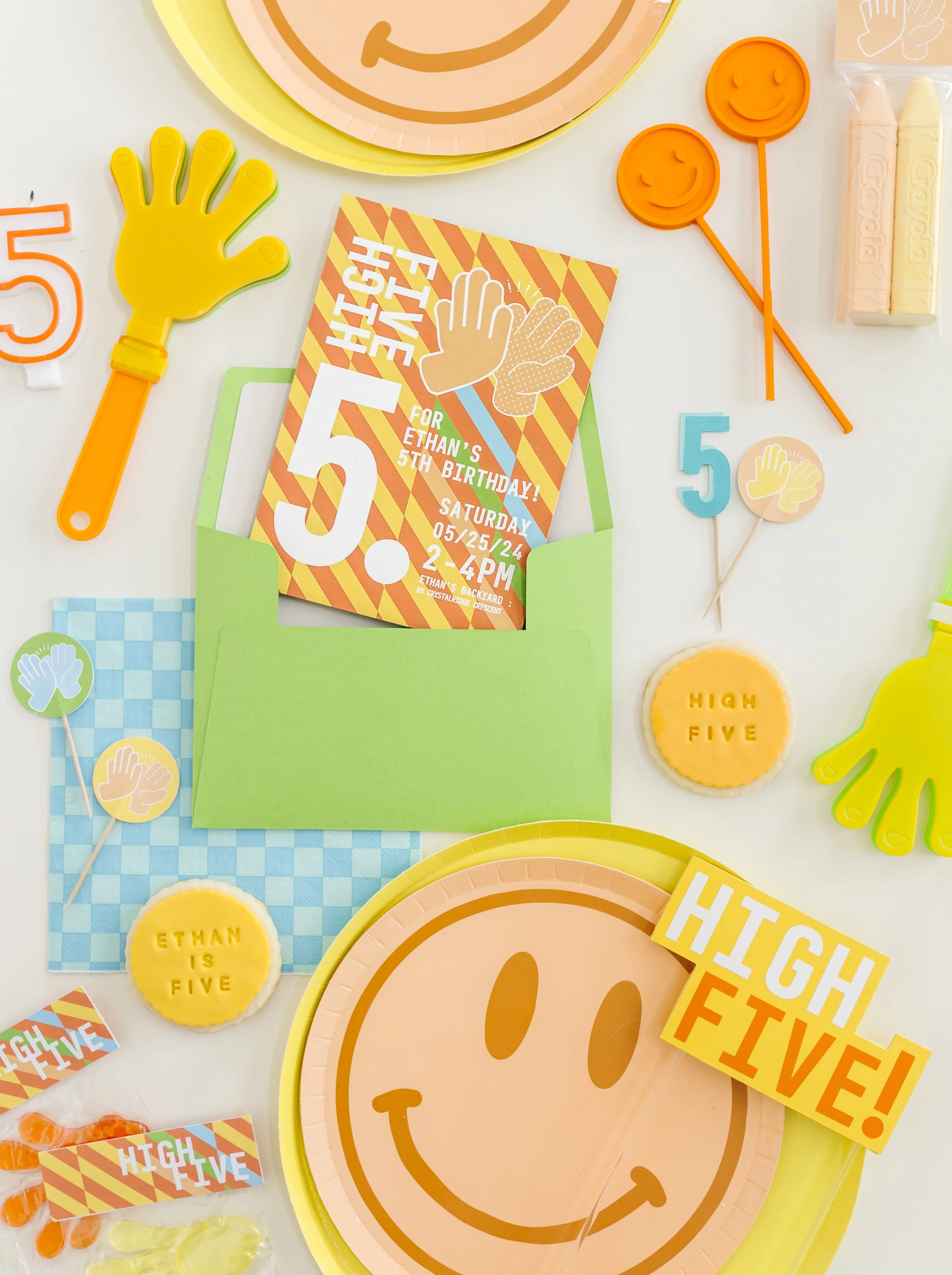 Colorful birthday party table with smiley face plates, high five hand clappers, decorated cookies with 'Ethan is five' and 'High Five!' text, birthday invitation, and party favors in bright colors.