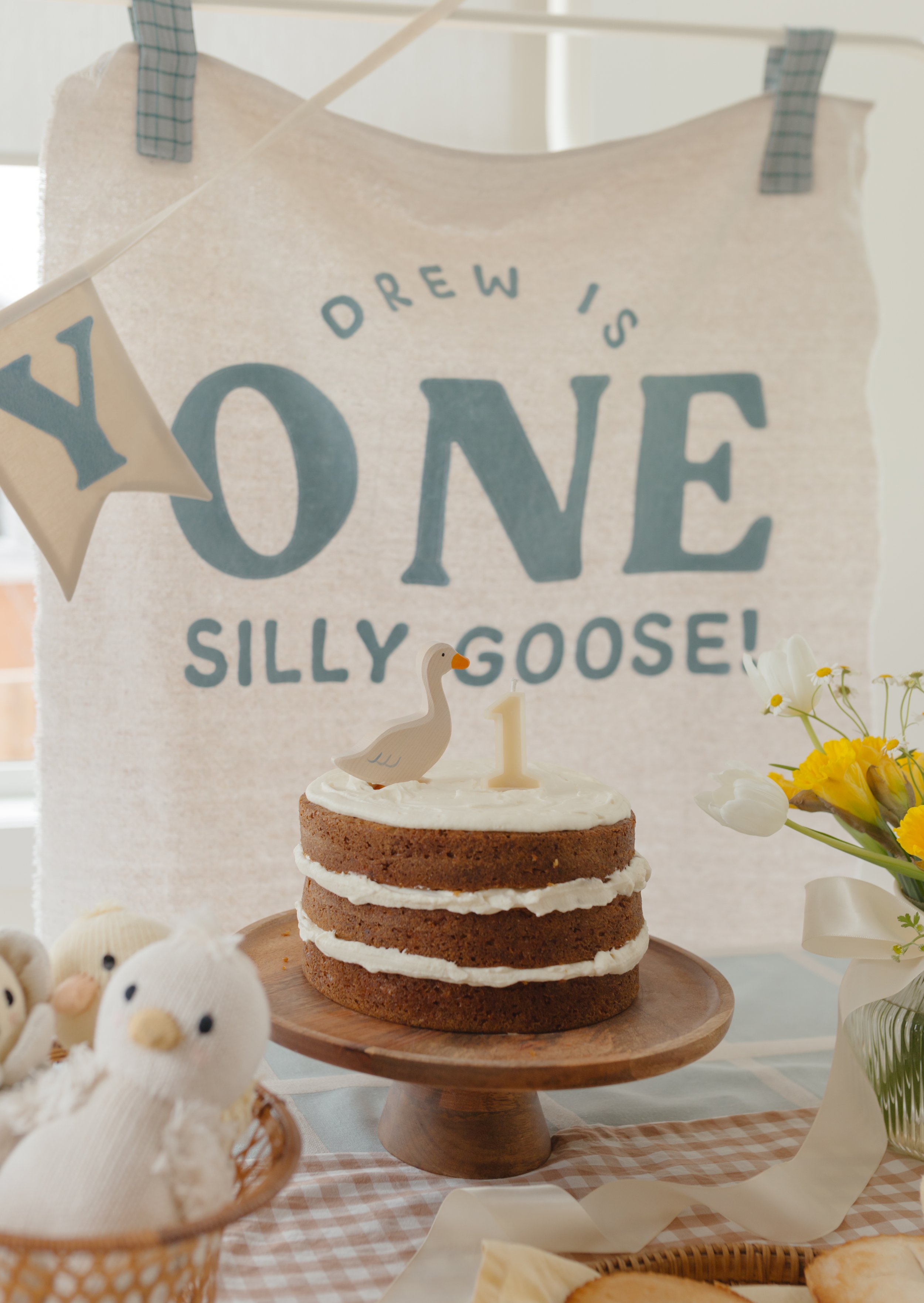 A birthday table setup with a small layered cake topped with a goose figurine and the number 1 candle, surrounded by plush duck toys and a flower arrangement. In the background, a sign says "Drew is one, silly goose!" with a banner spelling "Yay".