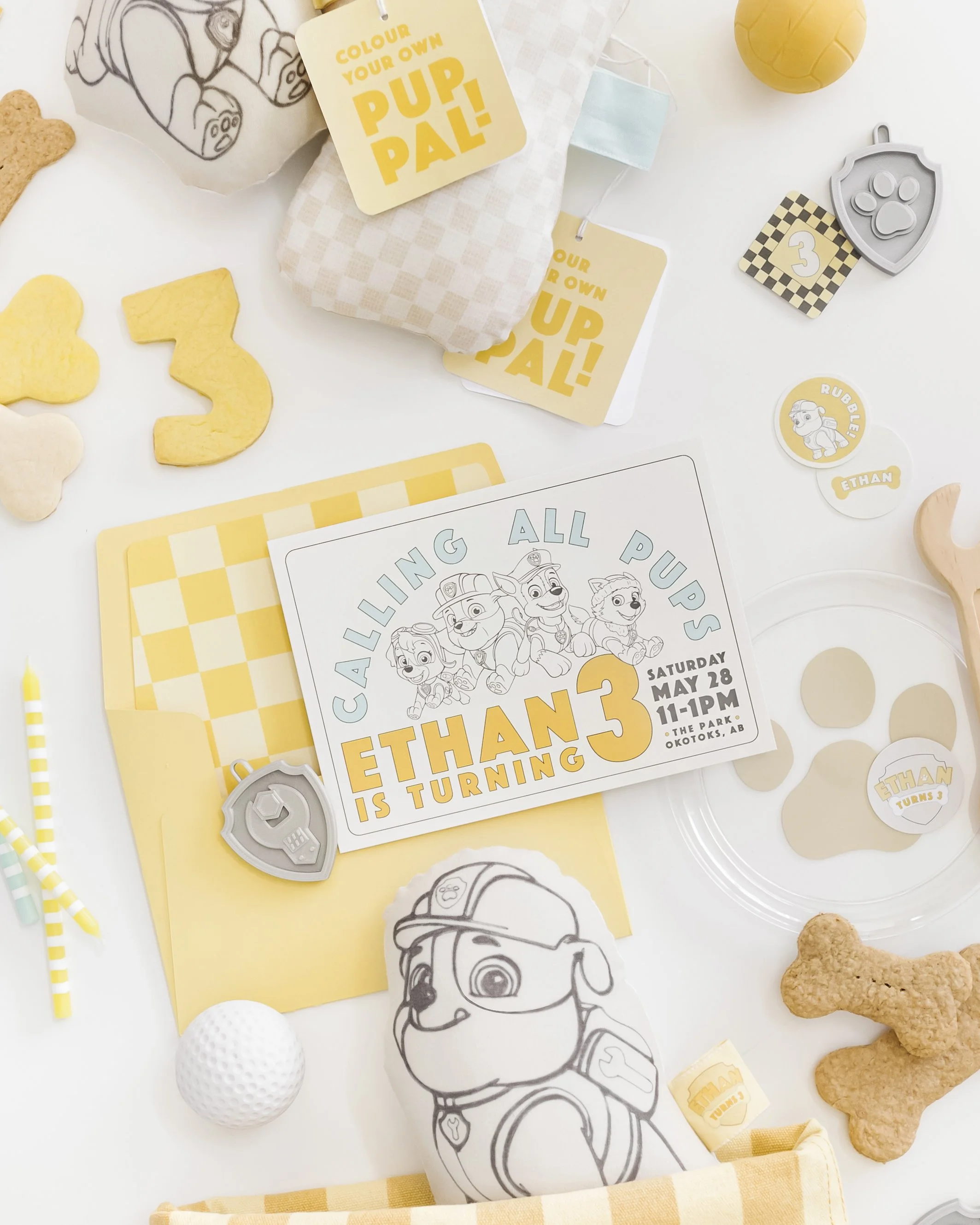 Dog themed birthday party invitation and decorations, including cookie and paper cutouts of dogs, a golf ball, dog paw print stickers, and dog character stickers, with a yellow, white, and gray color scheme.