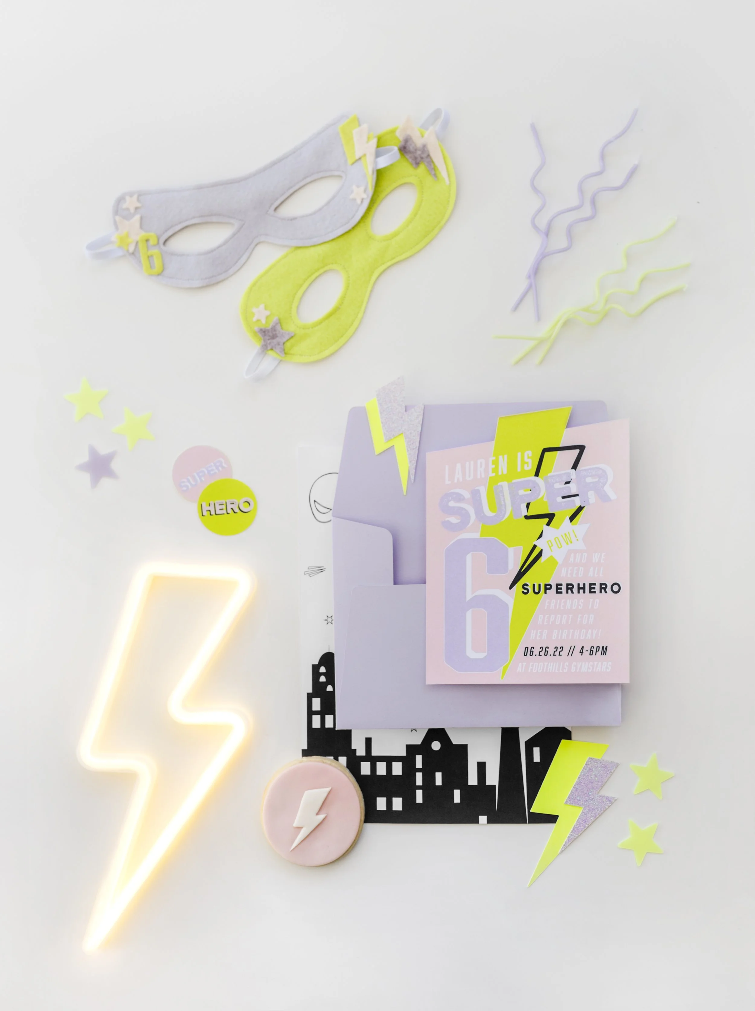 A superhero-themed birthday invitation surrounded by glow-in-the-dark masks, star and lightning decorations, and a neon lightning bolt light.