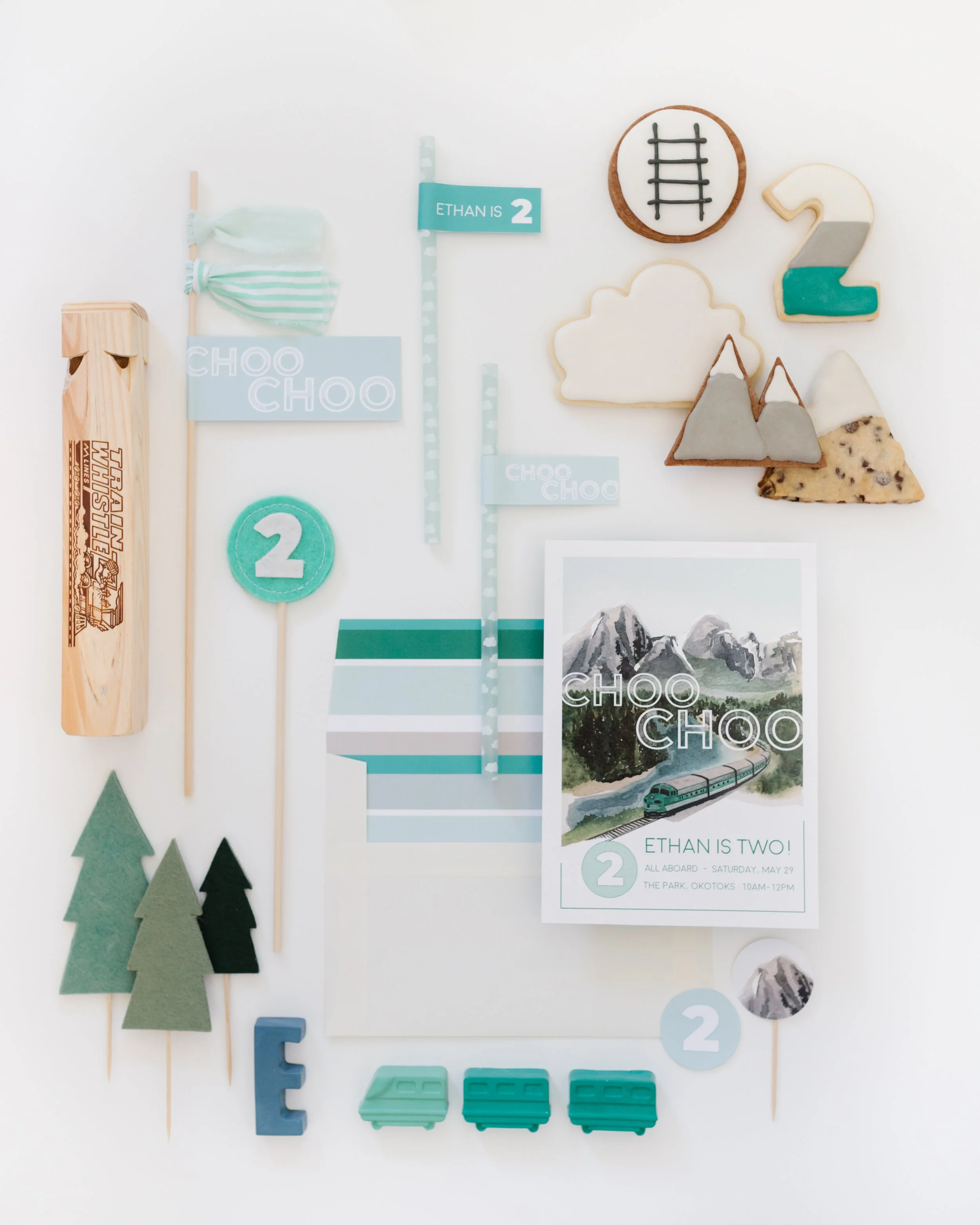 Collection of items for a children's birthday party with a train theme, including cookies, paper flags, felt trees, a train ticket, and train-shaped candies, all in green and blue tones.