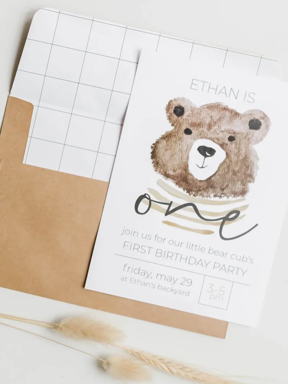Invitation card for a first birthday party featuring a watercolor illustration of a bear's face and the word 'love.' The event details specify the date as Friday, May 29, at Ethan's backyard, for children aged 3-5 years old. The invitation is partially inside a brown envelope, with some decorative dried grass in the bottom left corner.