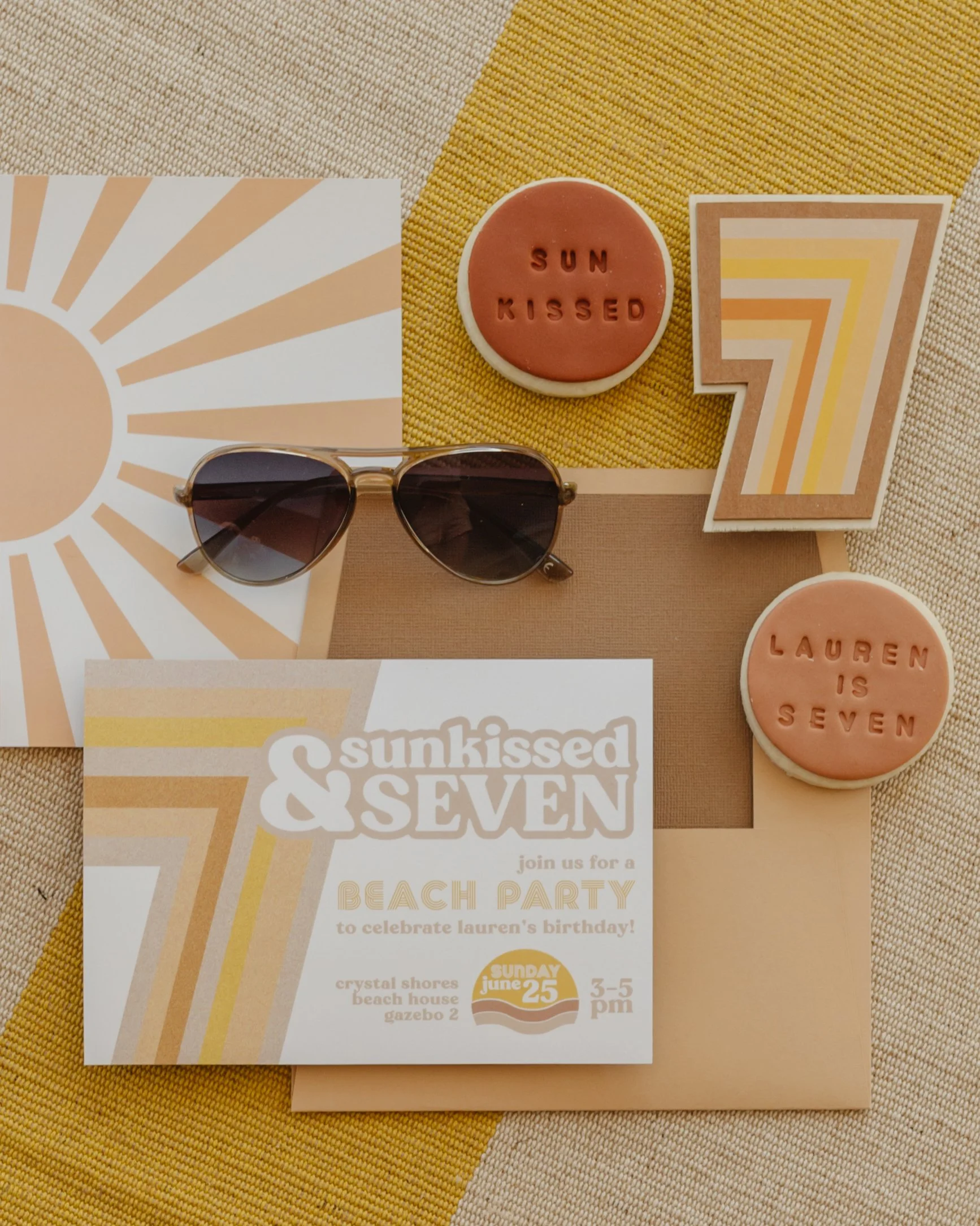Beach-themed birthday party invitation with sunglasses, two pink cookies engraved with 'SUN KISSED' and 'LAUREN IS SEVEN,' a retro-style notebook, and a decorative card with the number seven, all set on a yellow and beige checkered background.