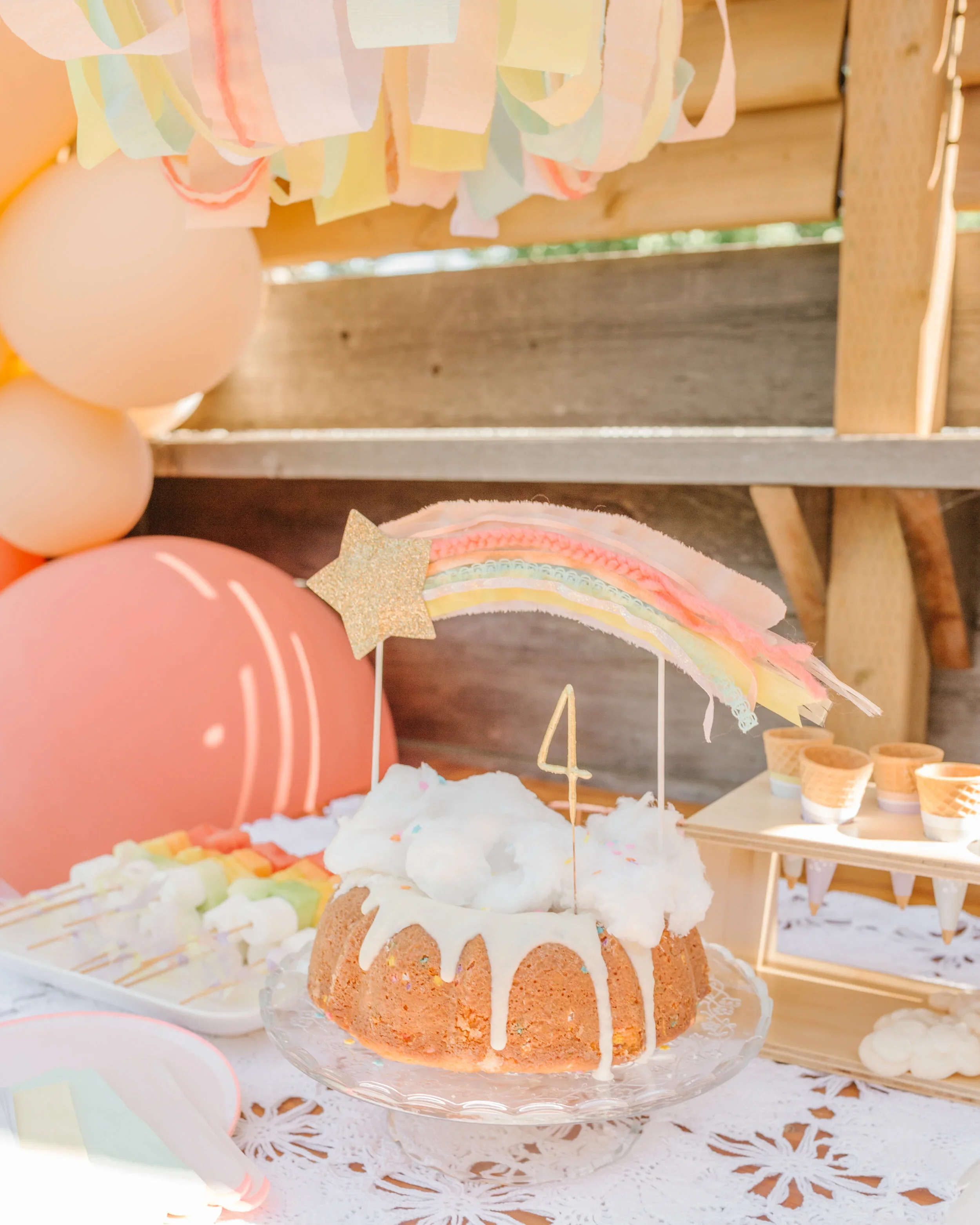 A birthday party table decorated with pastel balloons and a cake topped with whipped cream, a rainbow decoration, and a number 4 candle.