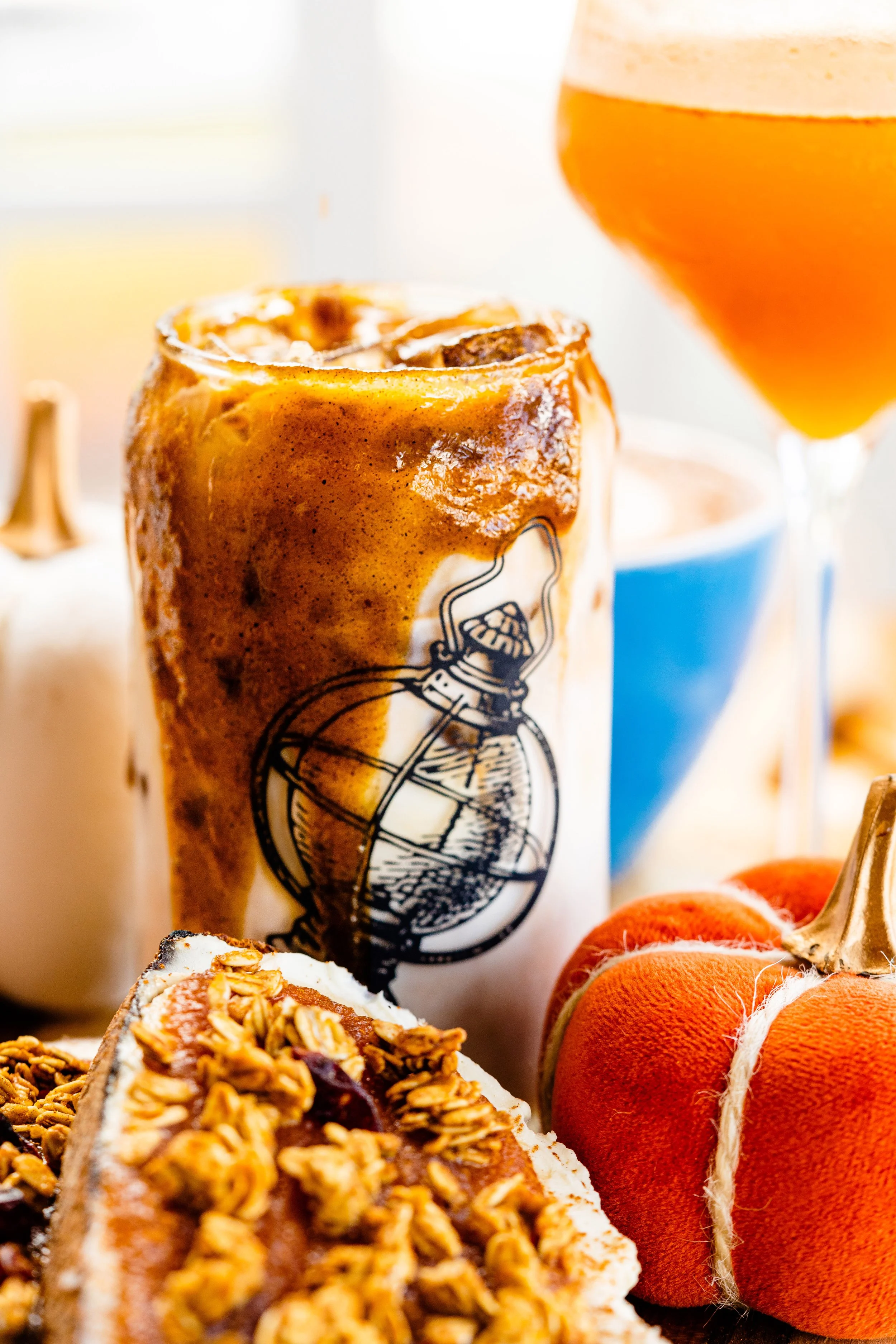 Close-up of a glass of beer topped with foam, a cinnamon roll with icing, a slice of granola bar, orange decorative pumpkins, and a blue bowl in the background.