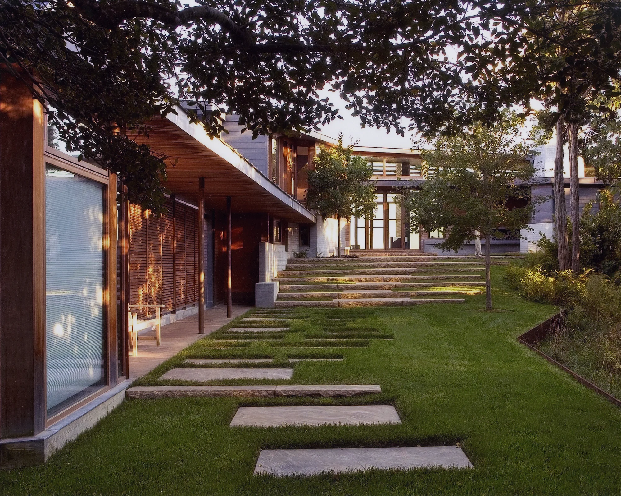 This site is situated in a dense coastal woodland on a dramatic bluff overlooking Pleasant Bay and the Atlantic Ocean. The gravel drive weaves through the woodland and gently slopes down to arrive at an open landscape of rolling meadow dotted with groves of American Holly, Eastern Red Cedar, and a heritage Weeping Willow. The house, breezeway, and outbuildings are carefully sited to rise and fall with the undulating landscape. Following the breezeway, a path leads to a set of grass and granite steps that emphasize the natural topography at the house. The main house is sited at the high point of the property and incorporates a long stone terrace extending along the oceanside to capture water views and benefit from on southern exposure. The terrace is defined by a poured concrete wall that gently steps from east to west to follow existing grade. To unify the project, the same materials are used both within the interior and exterior spaces. Embedded within the terrace is a reflecting basin that spills into a runnel that moves along the wall and empties into a basin filled with findings from the beach below. Collaborators Charles Rose Architects Photography Charles Mayer