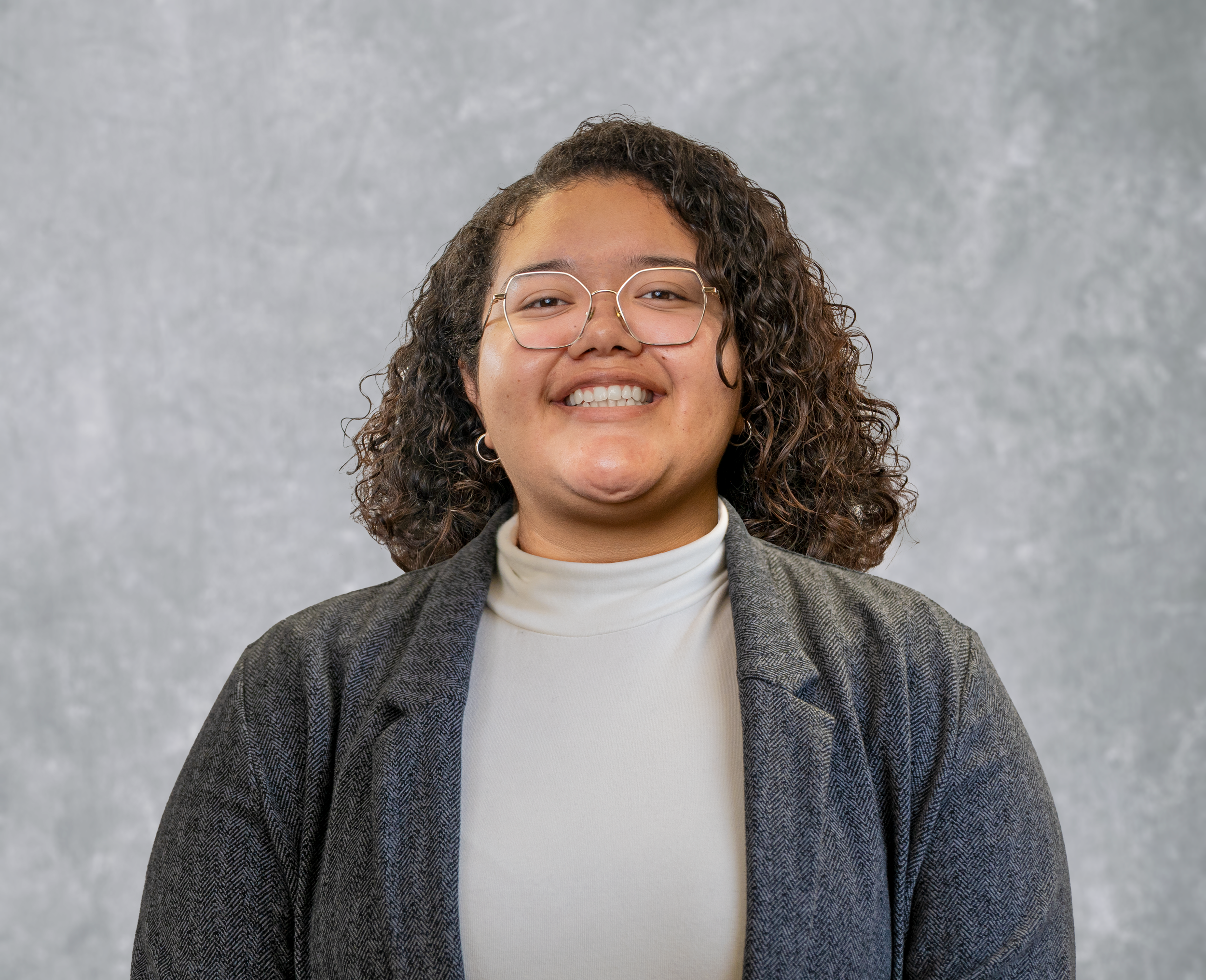 Staff member Nadia with curly hair, glasses, and hoop earrings smiling in front of a gray textured background, wearing a white turtleneck and a gray blazer.