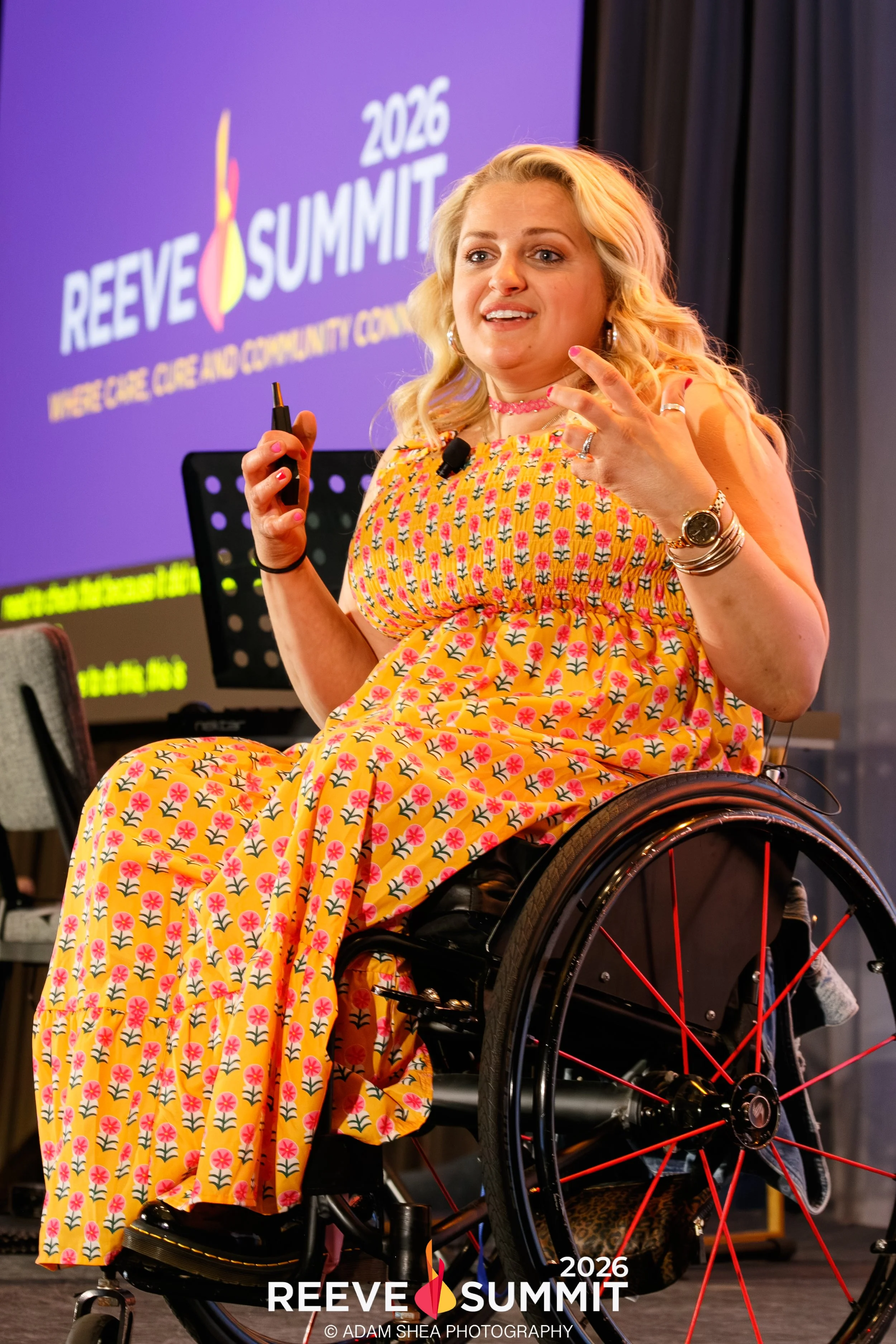 This is an image of the 2026 Reeve Summit Emcee, Ali Stroker. She is animatedly talking with her hands, wearing a yellow dress with little flowers, Doc Martens shoes, and she is seated in a manual wheelchair with red spokes on the wheels.