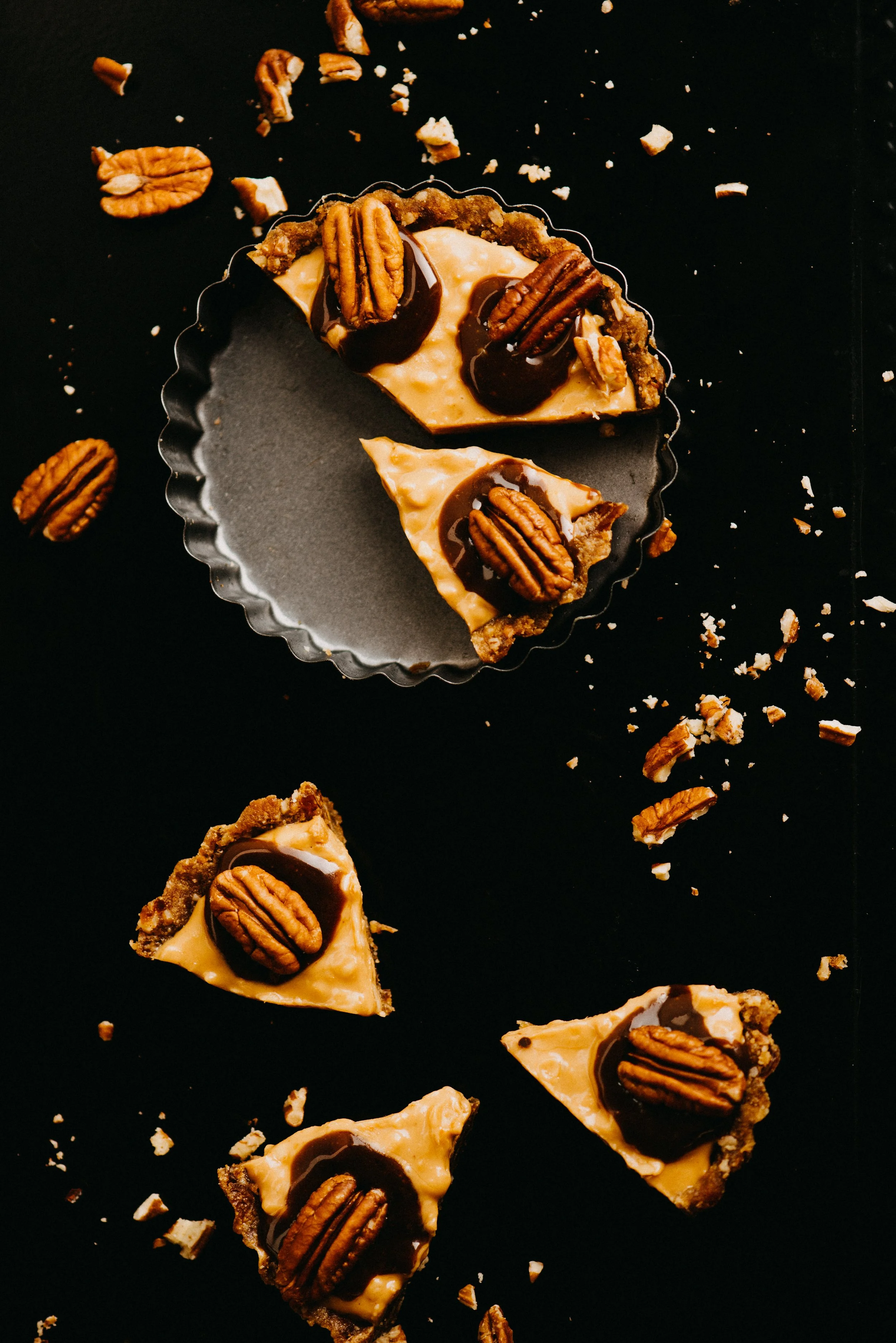 Pieces of pecan pie with chocolate and crushed pecans scattered on a black surface.