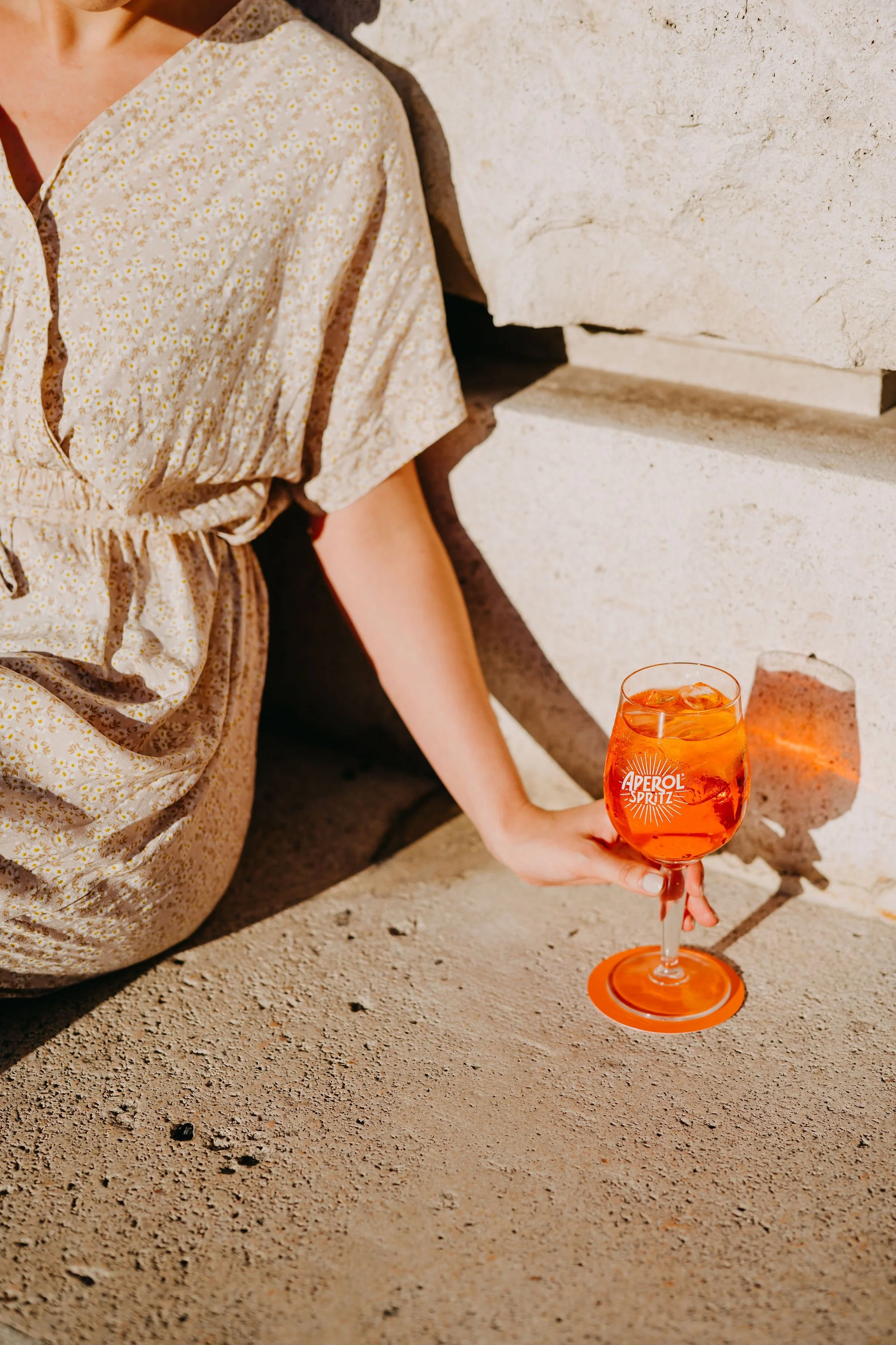 A woman sitting on the ground next to a stone wall, holding an orange-colored cocktail in a wine glass labeled 'Aperol Spritz' with ice cubes. She is wearing a light-colored, floral dress with short sleeves.