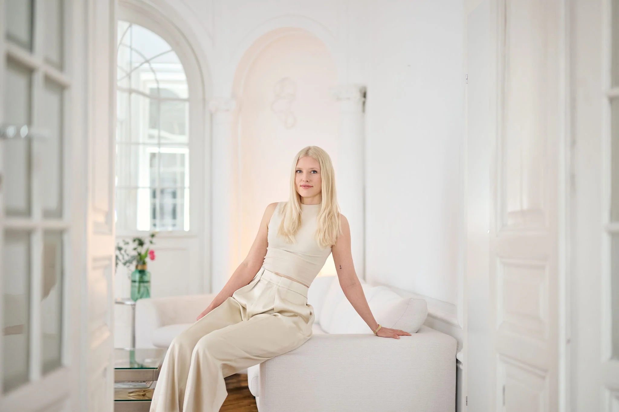 A woman with long blonde hair wearing a beige top and matching beige pants sitting on a white cushioned bench in a bright, airy room with large windows and white walls.