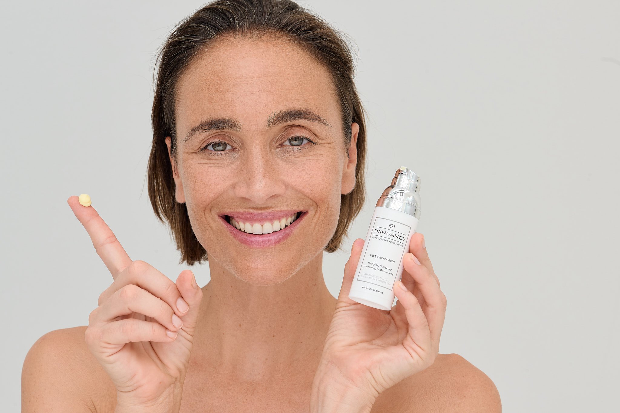 A woman with short brown hair and blue eyes smiling, holding a bottle of skincare product labeled 'Skinuance' and pointing to a small amount of cream on her finger.