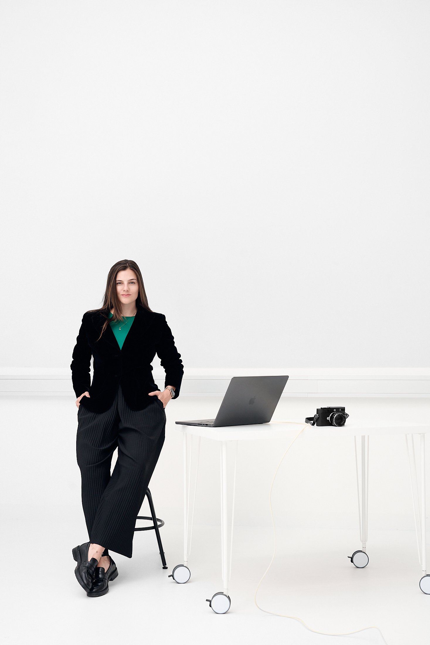 A woman standing in a bright, minimalistic office space with a white background, dressed in a black blazer, green top, and black striped pants, with her hands in her pockets. On the table behind her, there is a laptop, a camera, and a chair.