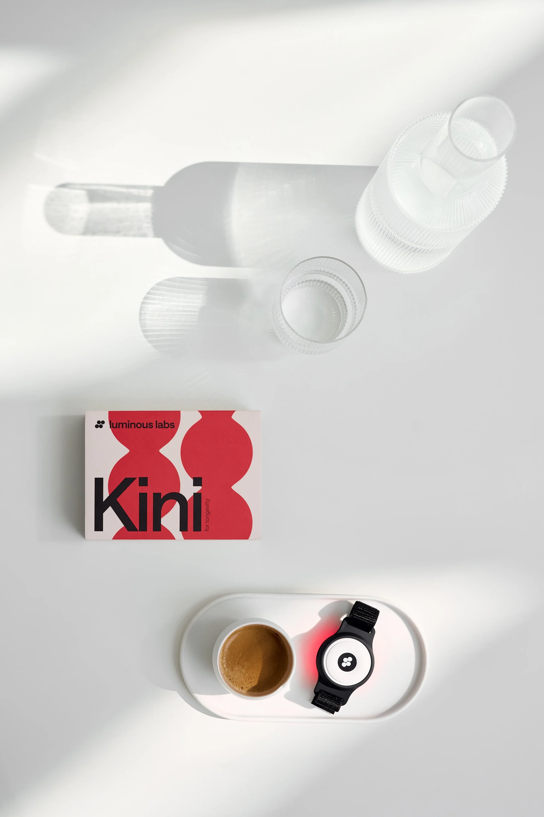 A flat lay of a white surface with a box of Luminous Labs Kini supplements, a small glass of water, a cup of coffee, and a smartwatch with a black strap. The shadows cast by the water glass create a pattern on the surface.