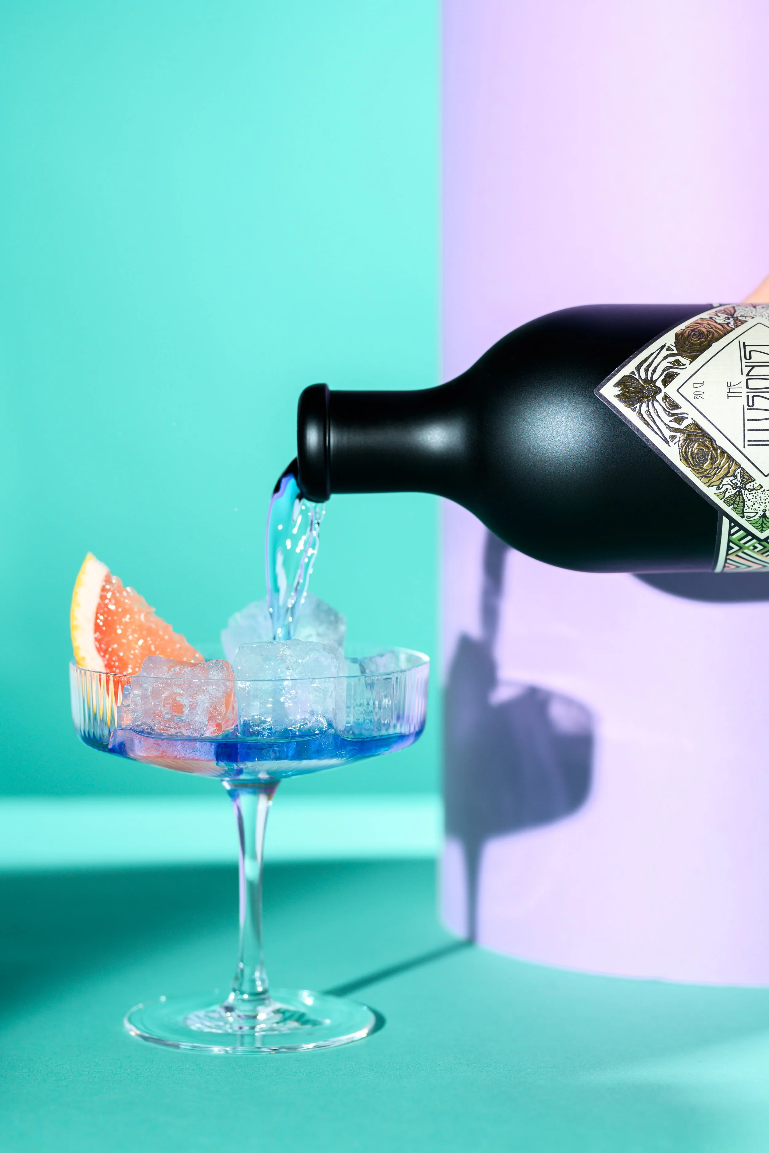 A black bottle pouring a clear liquid into a glass filled with ice and grapefruit slices. The background features a split pastel-colored backdrop with green on the left and lavender on the right.