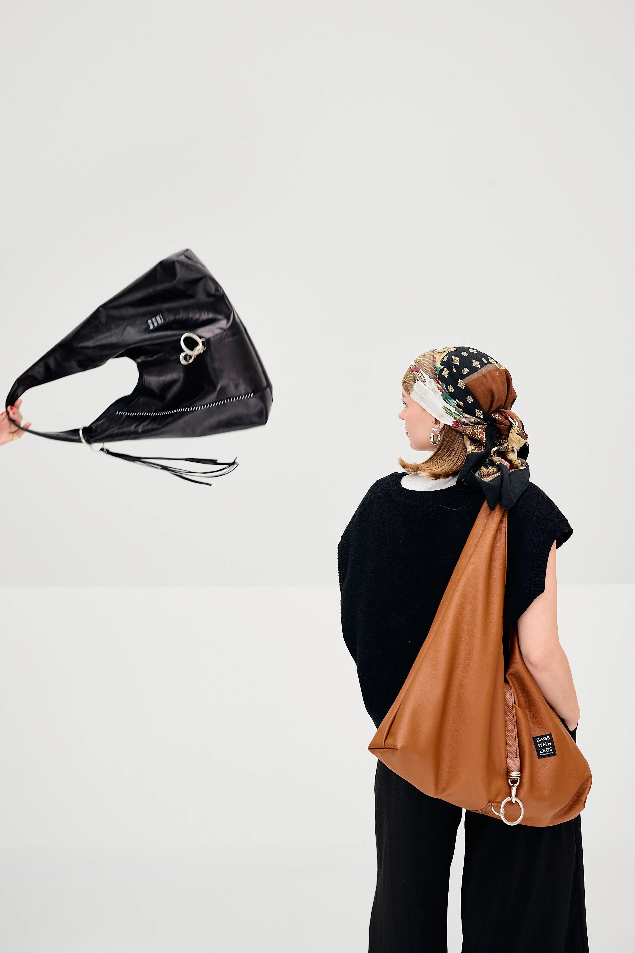A young woman with a floral headscarf and silver earrings wearing a black sleeveless top and black pants, carrying a large brown leather bag with a pin that says 'BAGS WITH LEGS', standing against a plain white background. An arm extending from the l