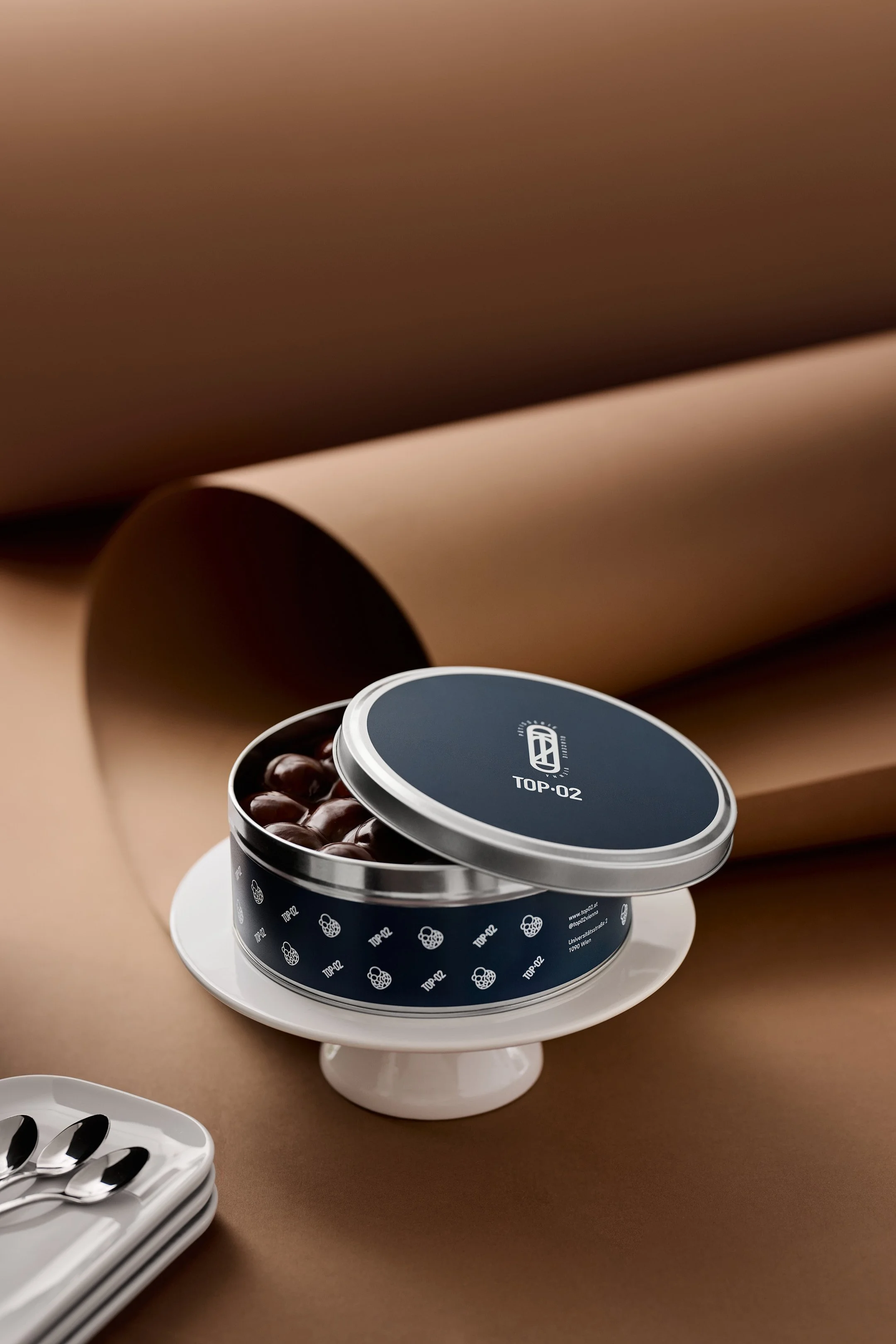 A round, black container of chocolates with a silver lid, labeled 'TOP-02', sitting on a white stand. In the background, there are brown papers or fabric and a white tray with silver spoons.