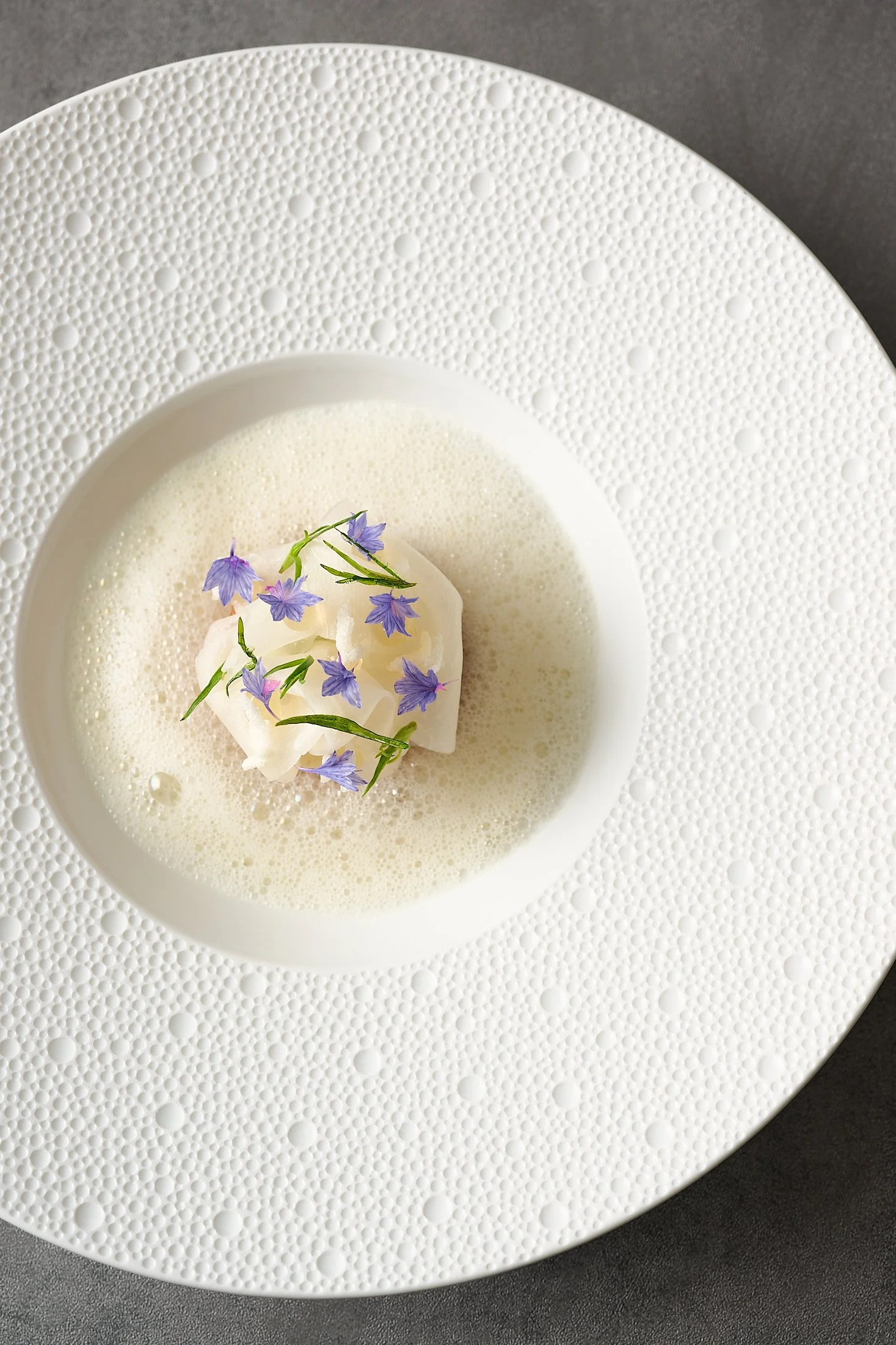 A white ceramic plate with textured dot patterns, holding a small serving of dessert topped with edible purple flowers and green herbs.