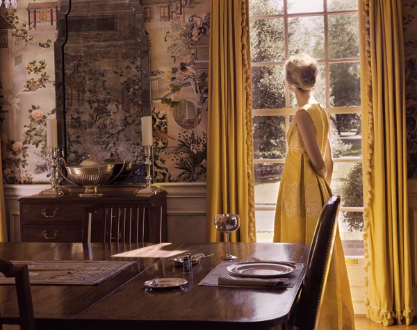 A woman looking out the window by Danielle Nelson-Mourning of a woman (herself) dressed in yellow dress looking out of a window with matching curtains in a dining room with ornate wallpaper.