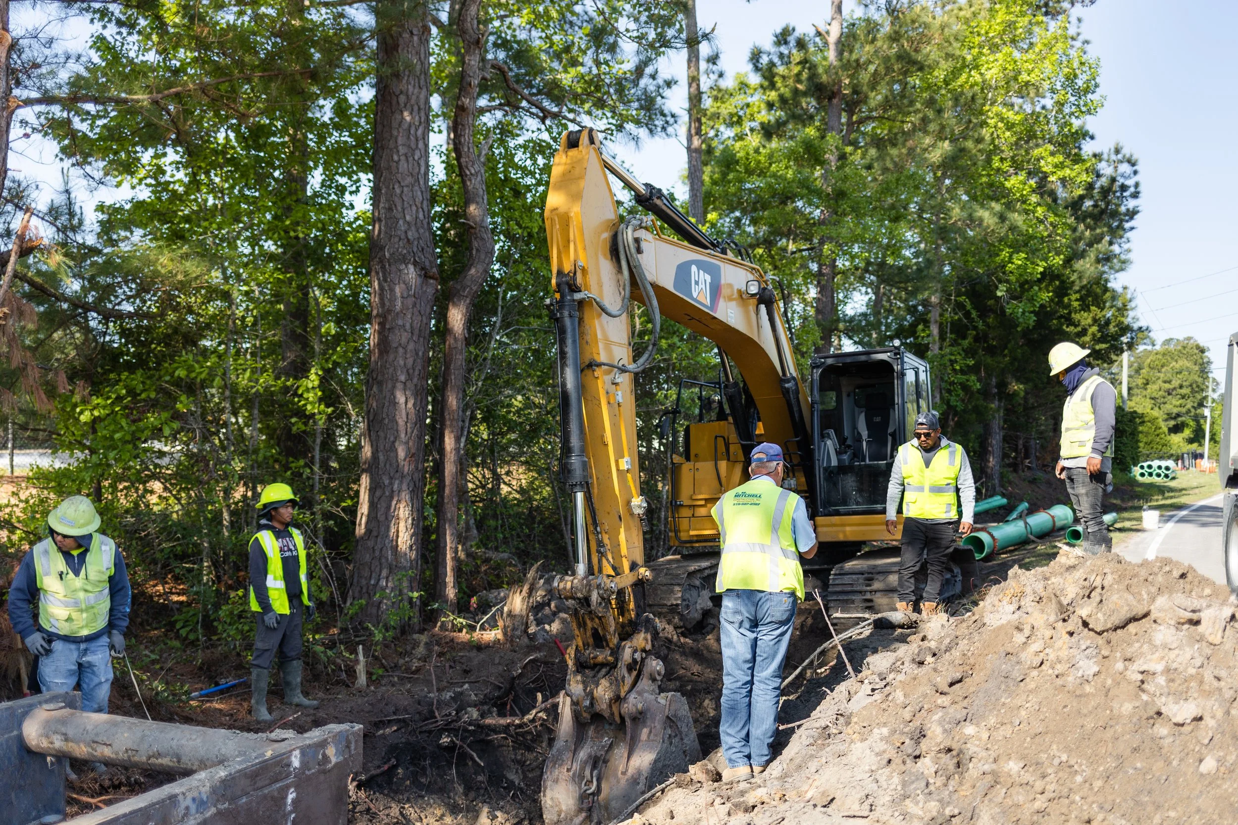 Construction workers and an excavator working on a road repair project surrounded by trees.