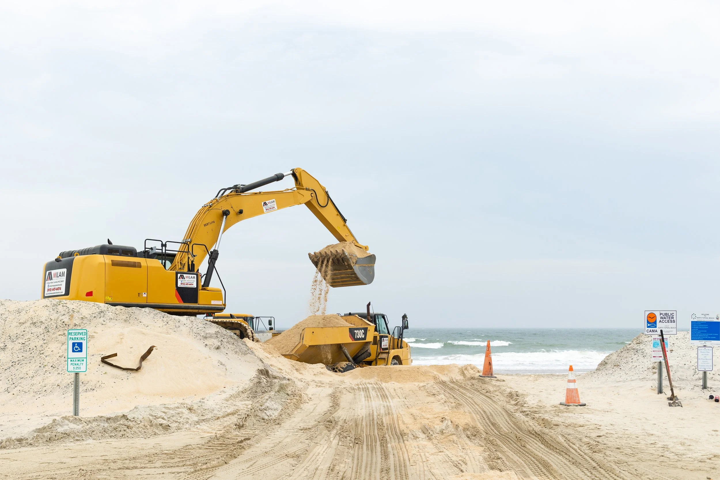Construction equipment on a sandy beach with signs indicating parking and water access, and orange traffic cones along the shoreline.