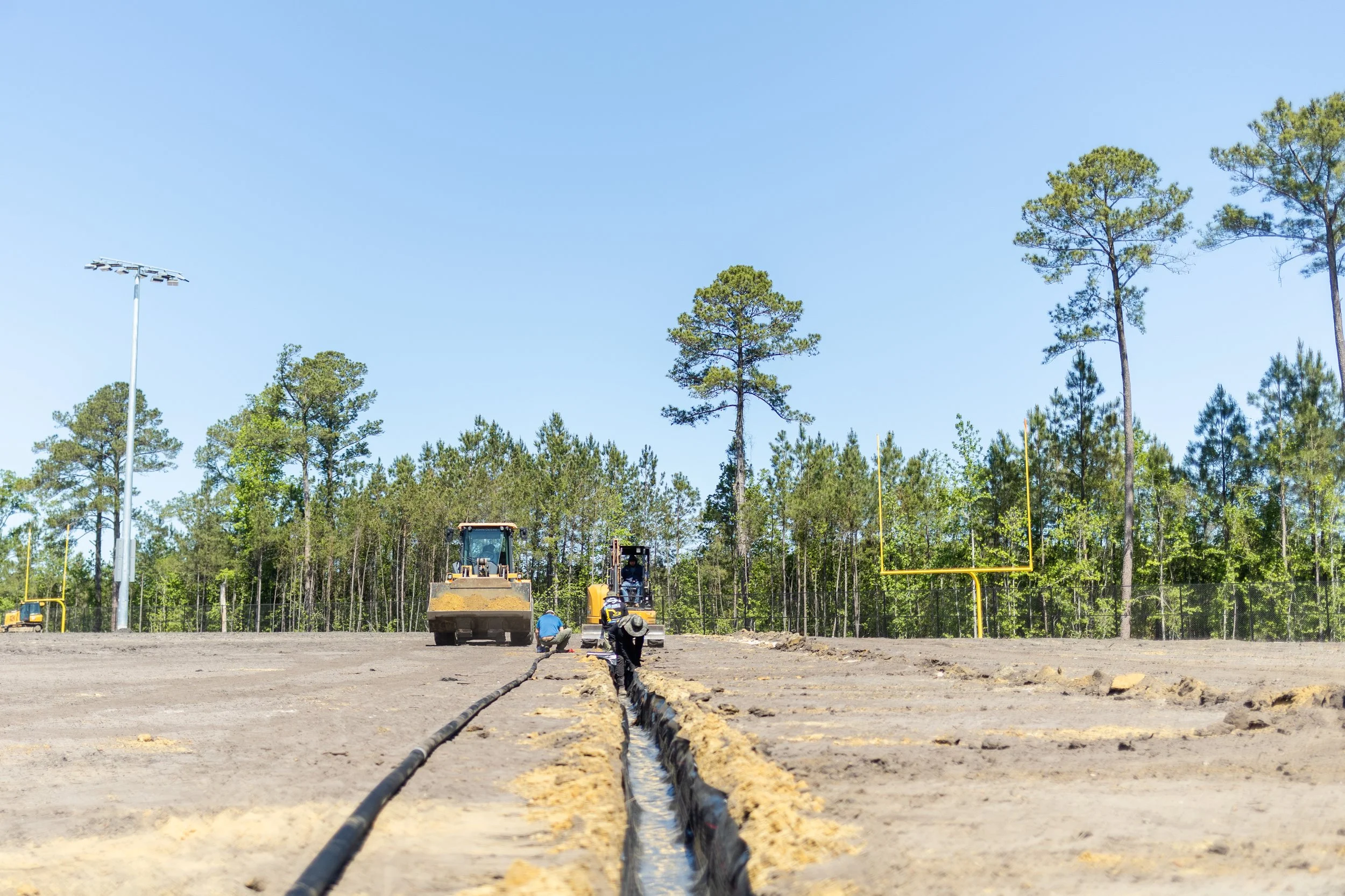 Construction workers leveling ground and installing piping on a baseball field, with tall trees and a clear blue sky in the background.