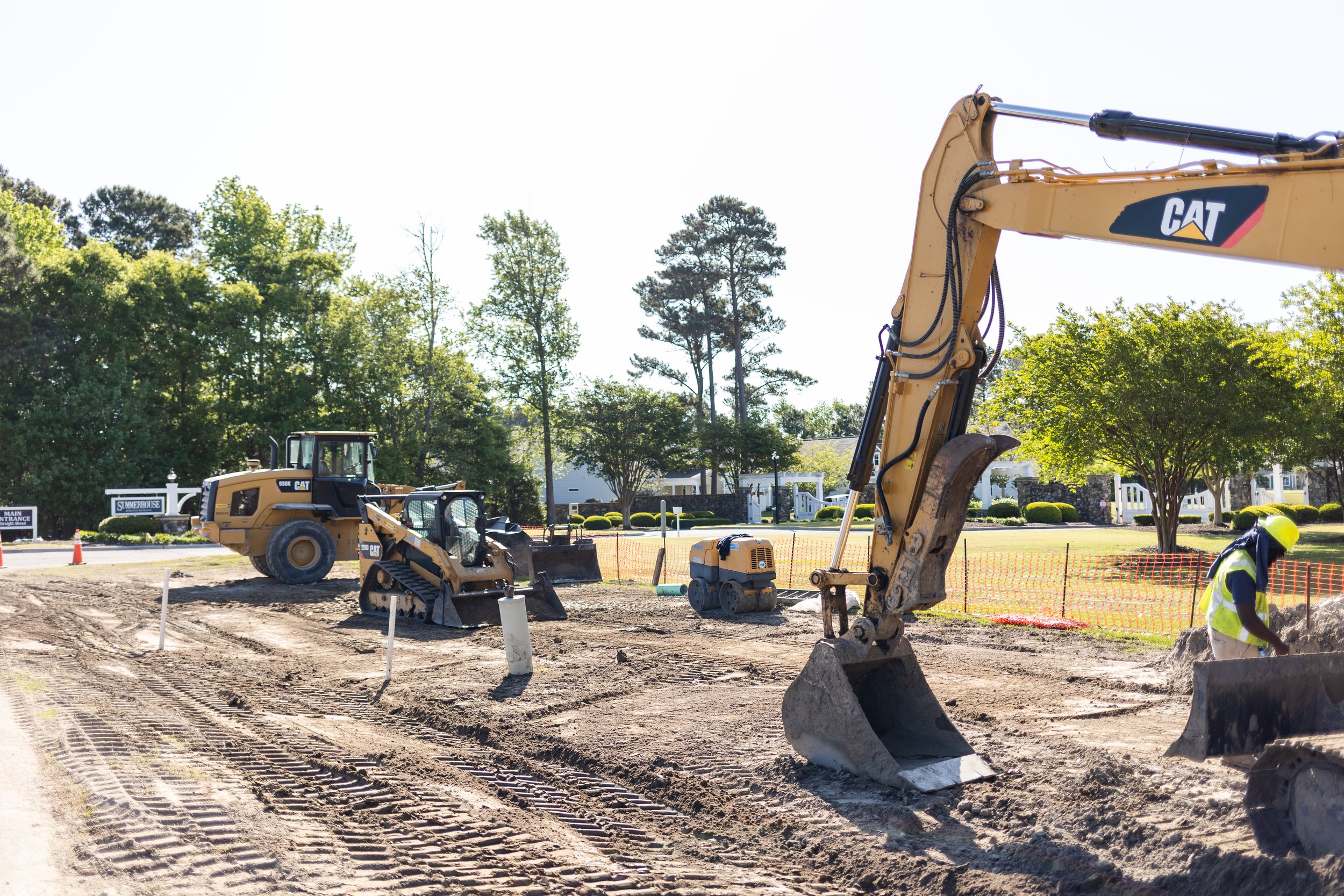 Construction site with machinery including a large excavator and a bulldozer, workers wearing hard hats, and a partly dirt and paved area with trees and a white fence in the background.