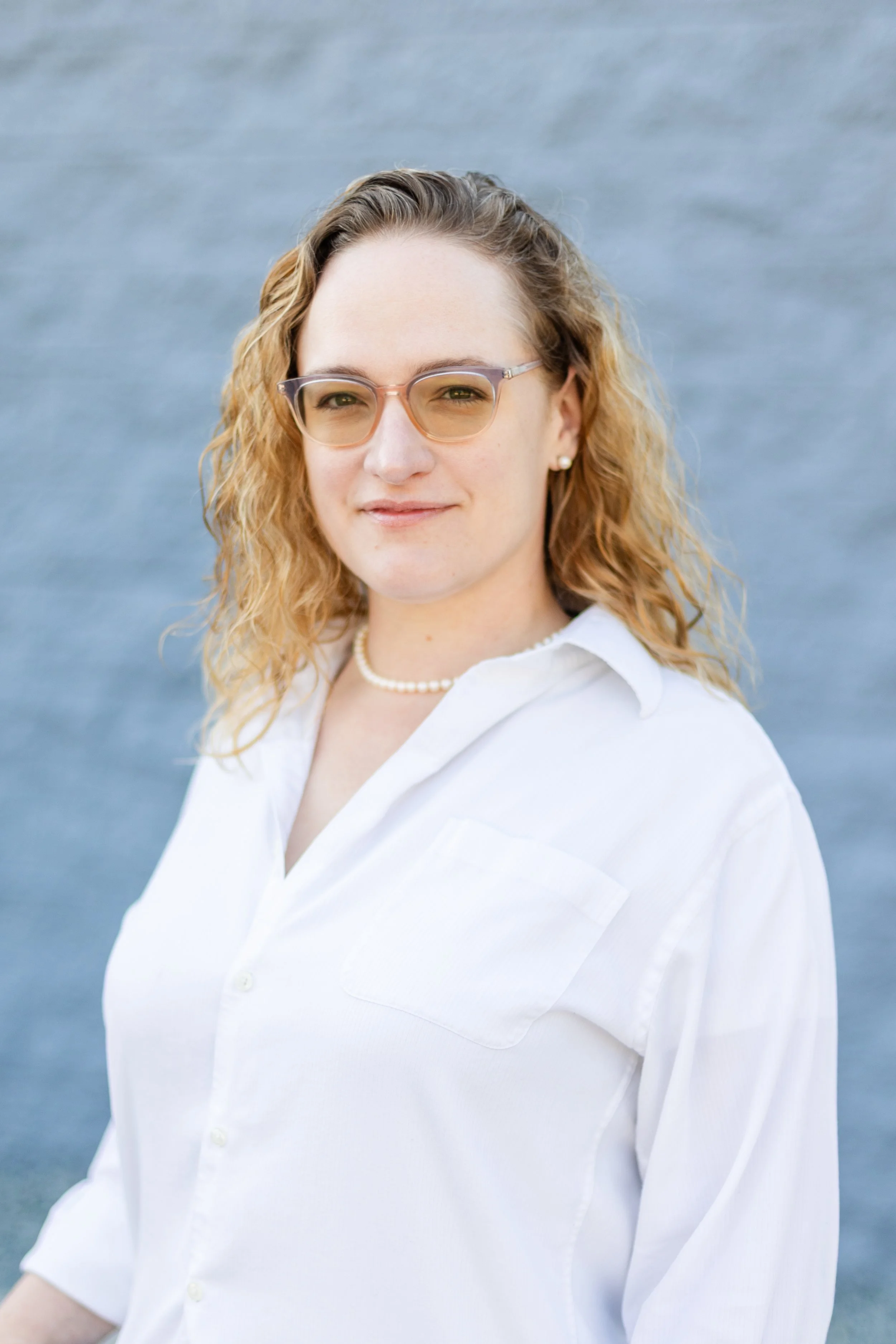 A woman with shoulder-length curly blonde hair, wearing clear glasses, a pearl necklace, earrings, and a white button-up shirt, standing in front of a body of water.