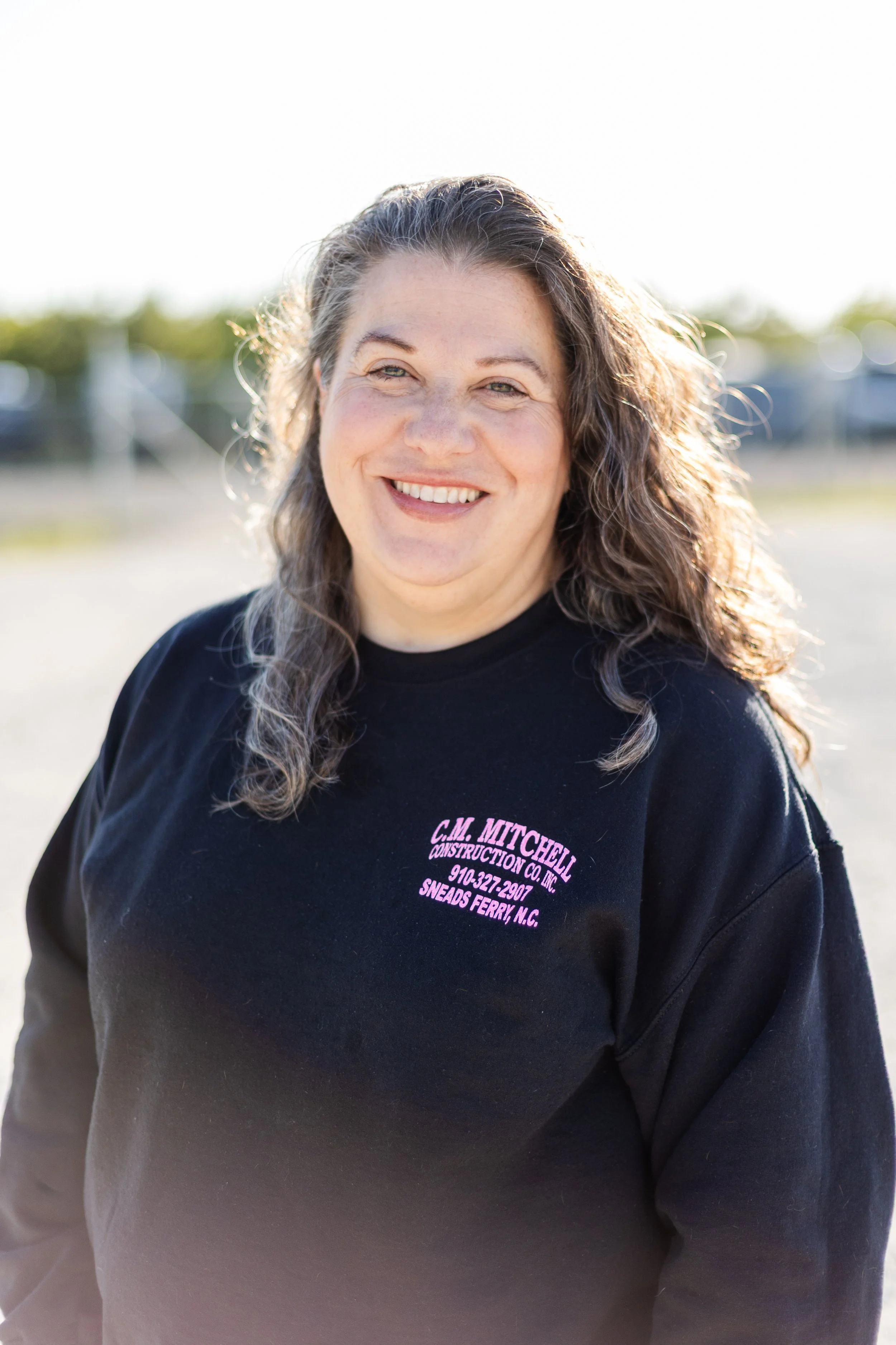 A smiling woman with long curly hair wearing a black shirt with pink text in an outdoor setting with blurred background.