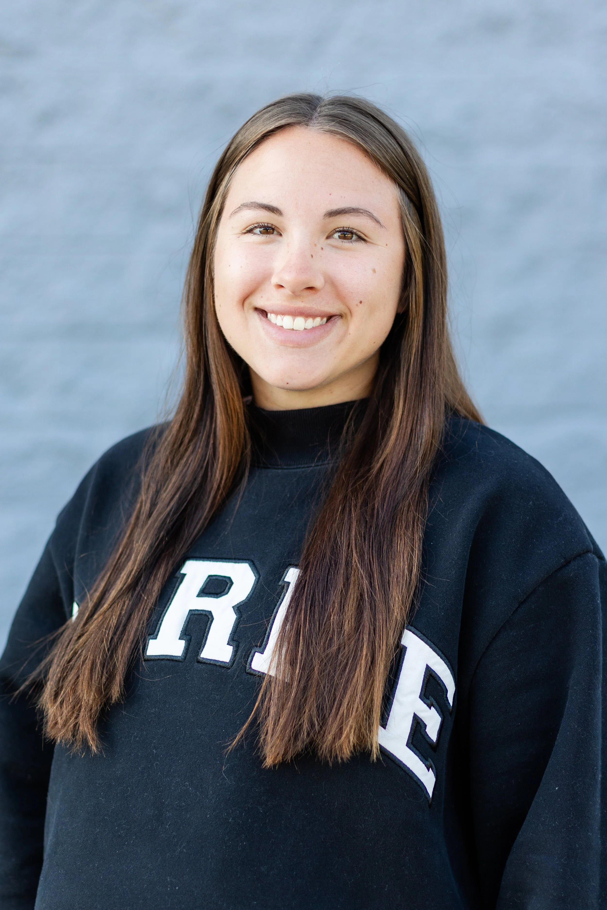 A young woman with long brown hair smiling, wearing a black sweatshirt with white letters, standing outdoors with a blurred water background.